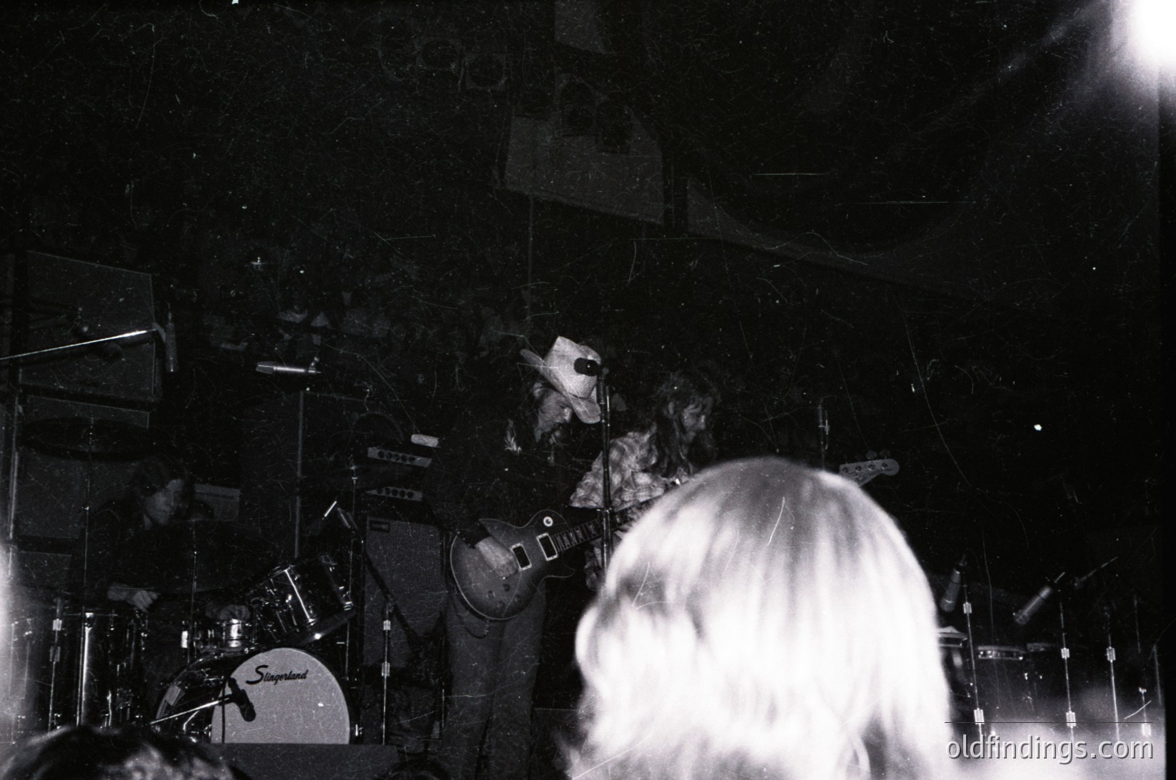 Black-and-white concert shot: guitarist playing electric guitar on stage with drum kit (Spergel brand) and amplifier. Crowd blurred in foreground. Likely 1970s–1980s rock/metal venue.