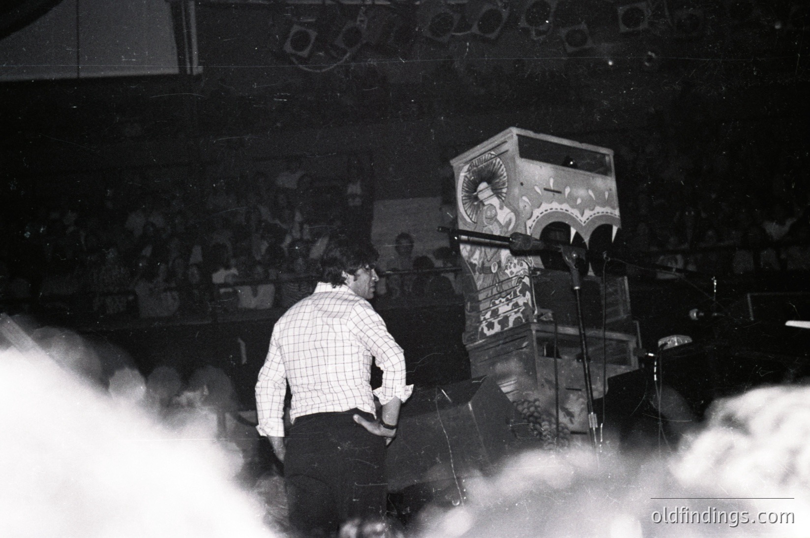 Vintage black-and-white concert shot of a performer on stage, back turned, wearing a checkered shirt and dark pants. Stage setup includes a vintage soda machine with "Coca-Cola" branding and a microphone stand. Crowd in background appears dense, suggesting a live music venue. Likely 1960s–1980s era.