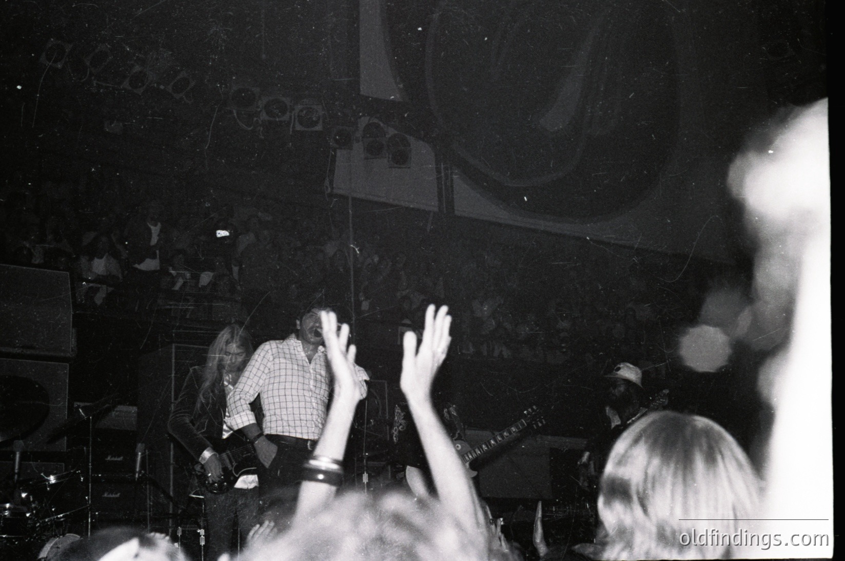Vintage black-and-white concert shot: performer on stage in checkered shirt, arms raised, with audience hands visible in foreground. Stage lighting and speakers suggest 1970s rock venue. Crowd appears engaged, capturing live music energy.