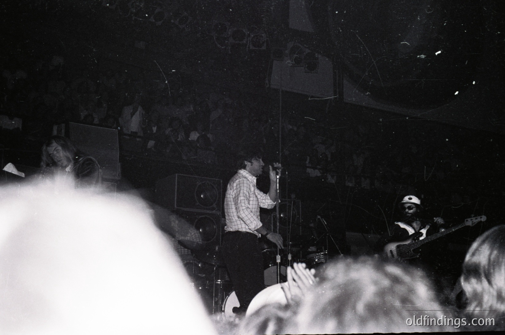 Black-and-white concert shot featuring a saxophonist and guitarist performing on stage, surrounded by a dense crowd. Stage lighting highlights instruments and performers against a dark backdrop. Crowd claps, suggesting live music event. Style and grain suggest mid-20th century (1950s–1970s) jazz or blues venue.