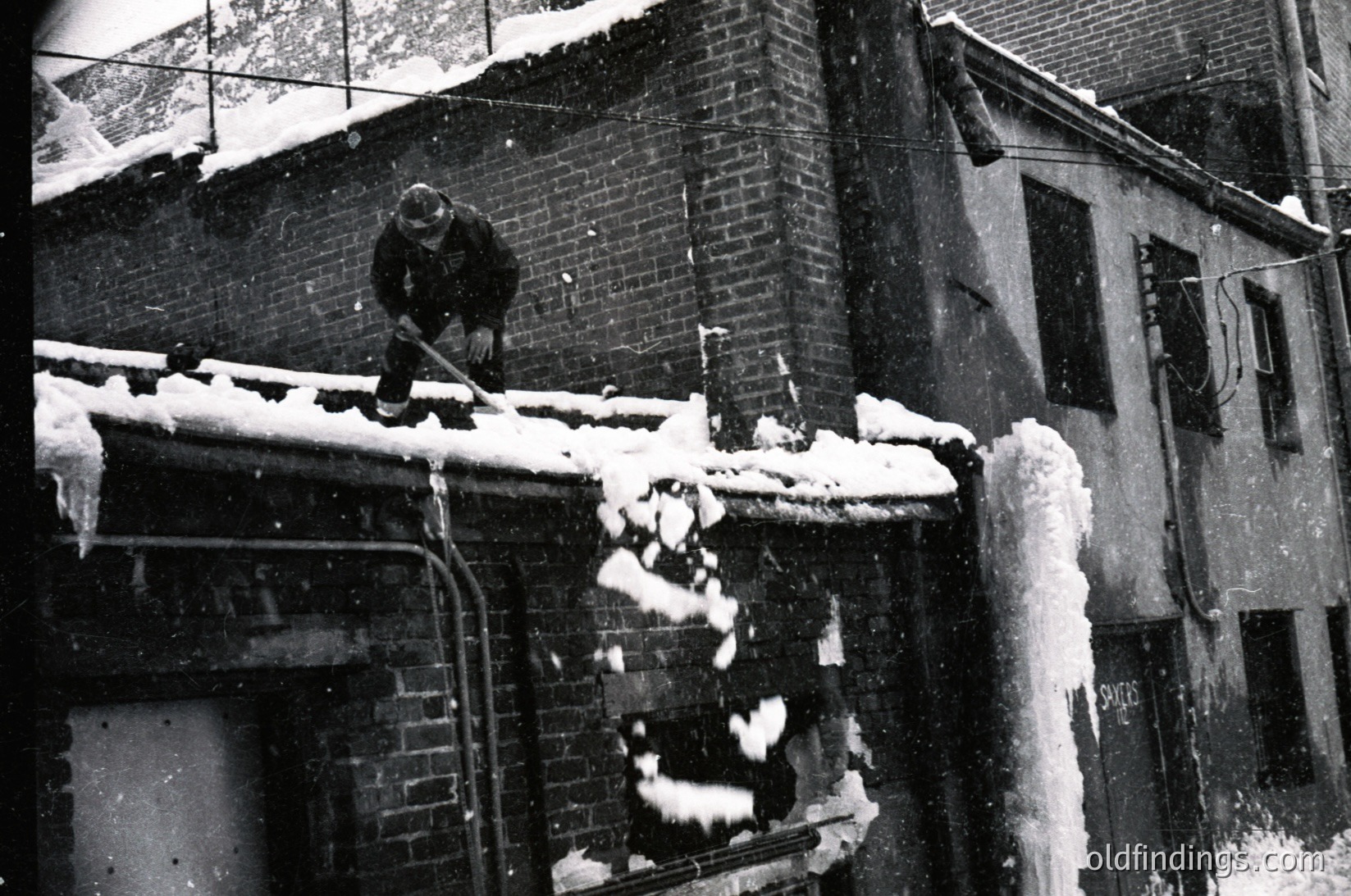 Black-and-white street scene: man clearing heavy snow from rooftop gutter using a shovel. Snow accumulates on brick buildings and railings. Mid-20th century urban winter setting.