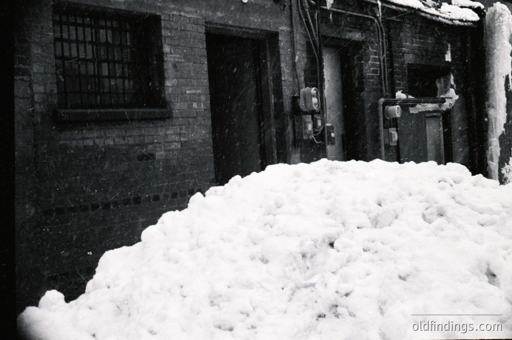 Mid-century industrial courtyard blanketed in deep snow, with brick walls and barred windows. Snowbanks obscure a fire escape and utility pipes. Classic black-and-white composition evokes 1950s–1960s urban winter.