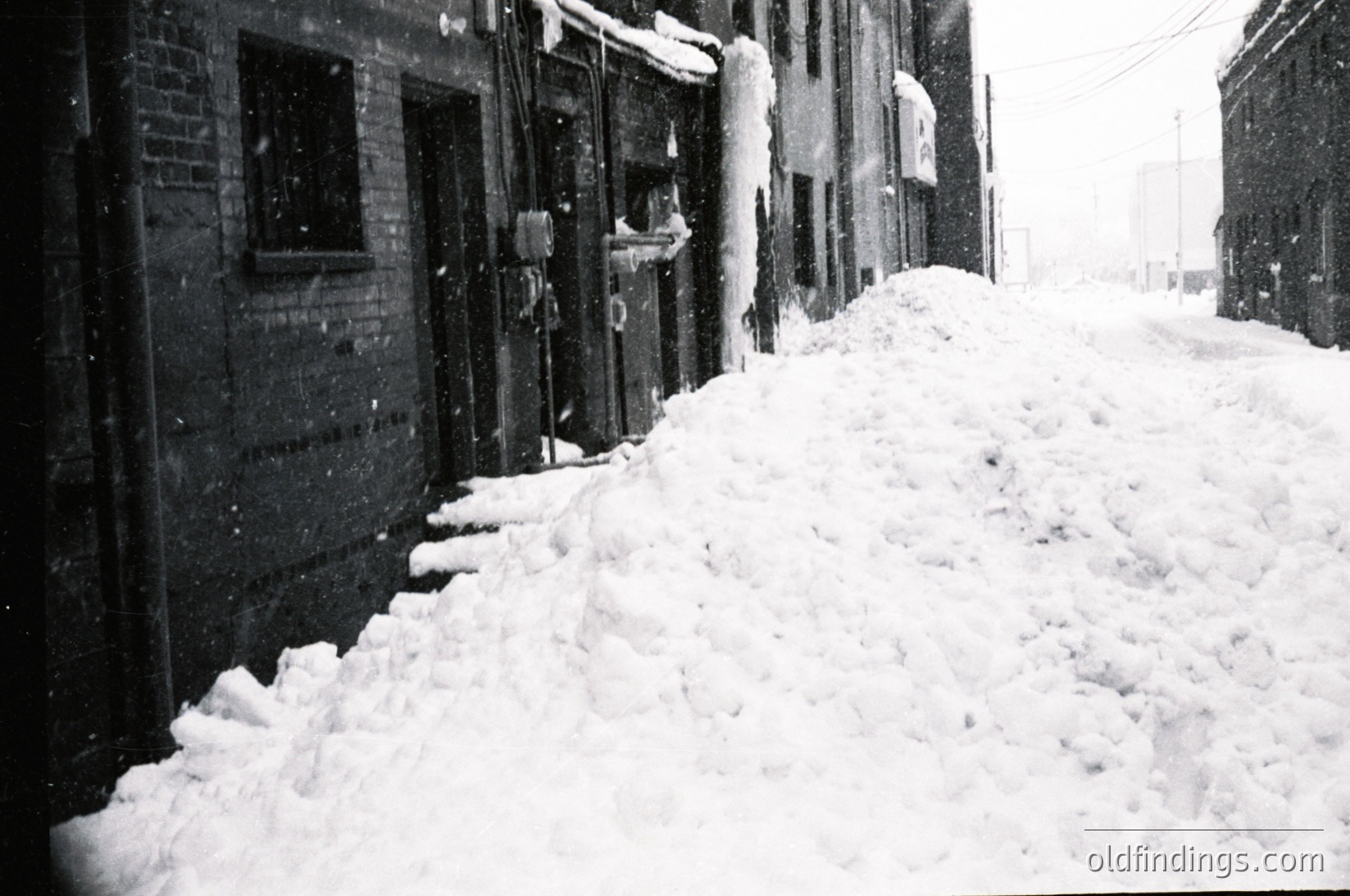 Snowdrifts pile high along a narrow alleyway flanked by brick buildings, likely residential or mixed-use. Heavy snowfall obscures doorways and windows, suggesting a mid-20th century urban setting. Black-and-white grain adds vintage aesthetic.