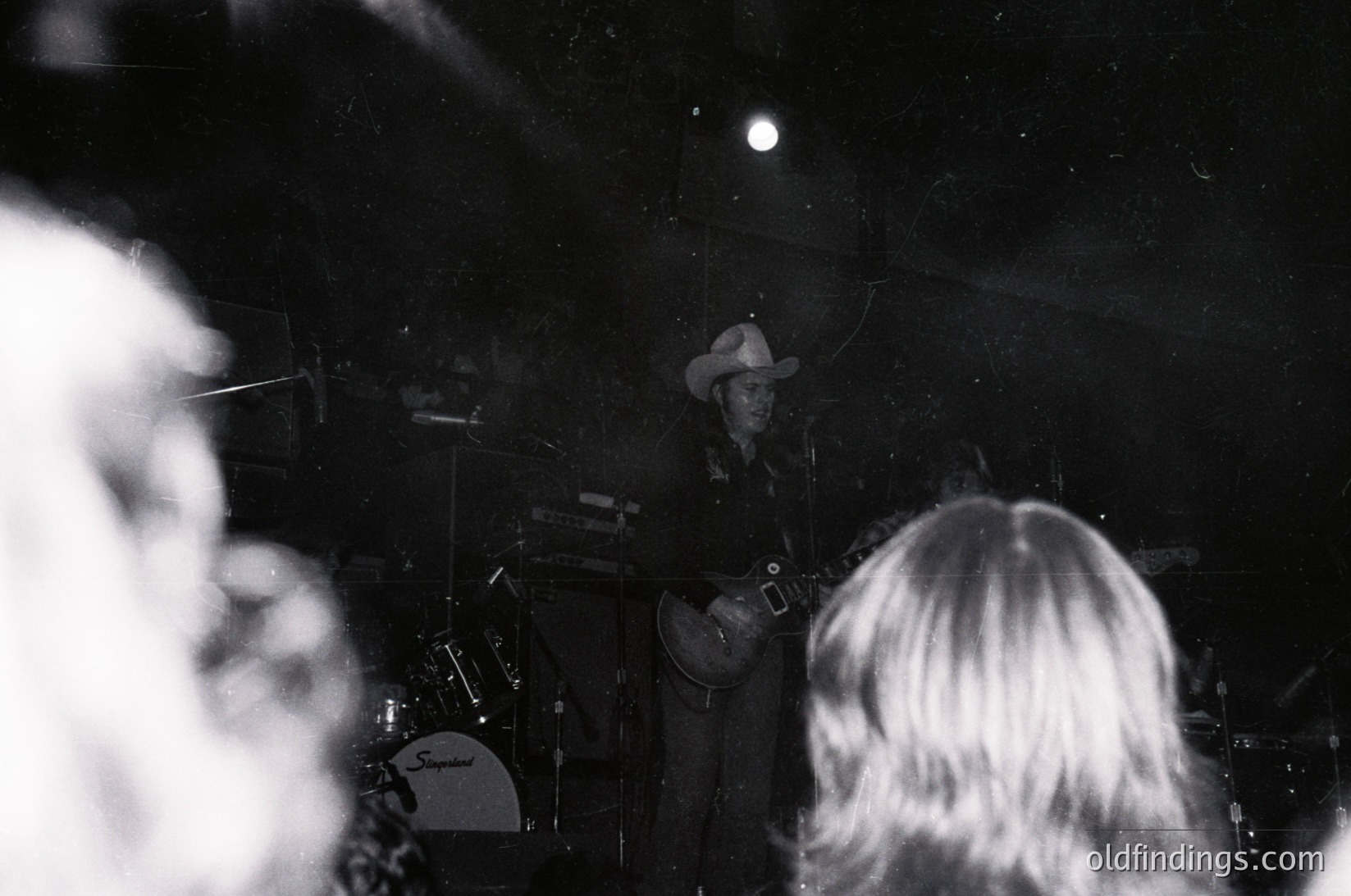 Vintage black-and-white concert shot: musician in cowboy hat playing guitar on stage, surrounded by foggy lighting. Audience silhouettes in foreground.