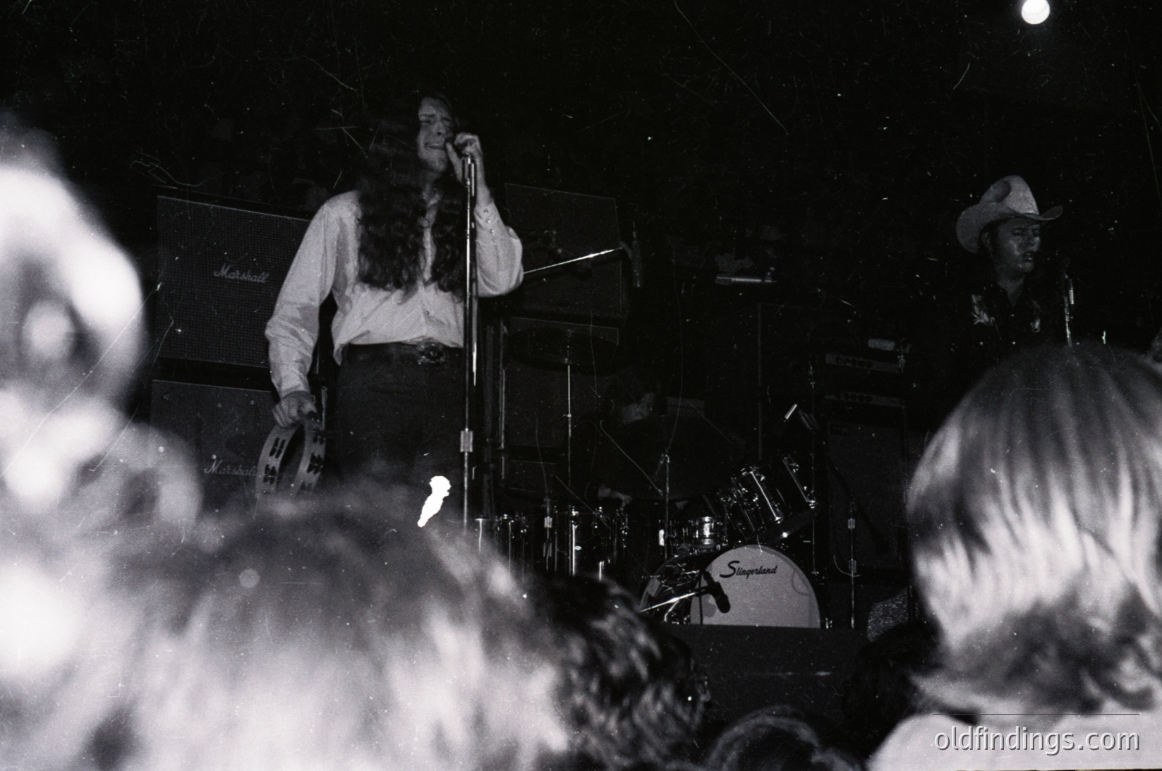 Vintage black-and-white concert shot of a band performing live, likely 1970s. Frontman in long hair and cowboy hat sings into mic, flanked by drummer (Suzuki cymbals) and guitarist. Crowd faces forward, silhouetted. Backlit stage equipment and partial signage ("Mallinson") visible. Authentic rock/music scene vibe.