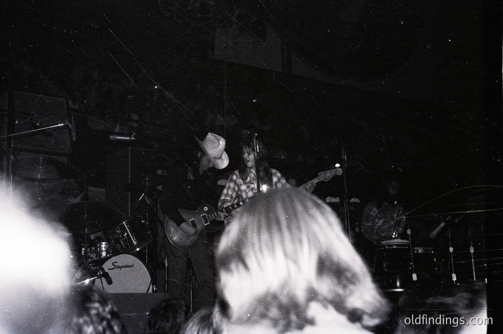 Black-and-white live band performance in dimly lit venue, featuring guitarist with checkered shirt and long hair playing electric guitar. Drum set (Spergel brand) and cymbals visible in background. Crowd silhouetted, suggesting mid-to-late 20th century rock concert atmosphere.