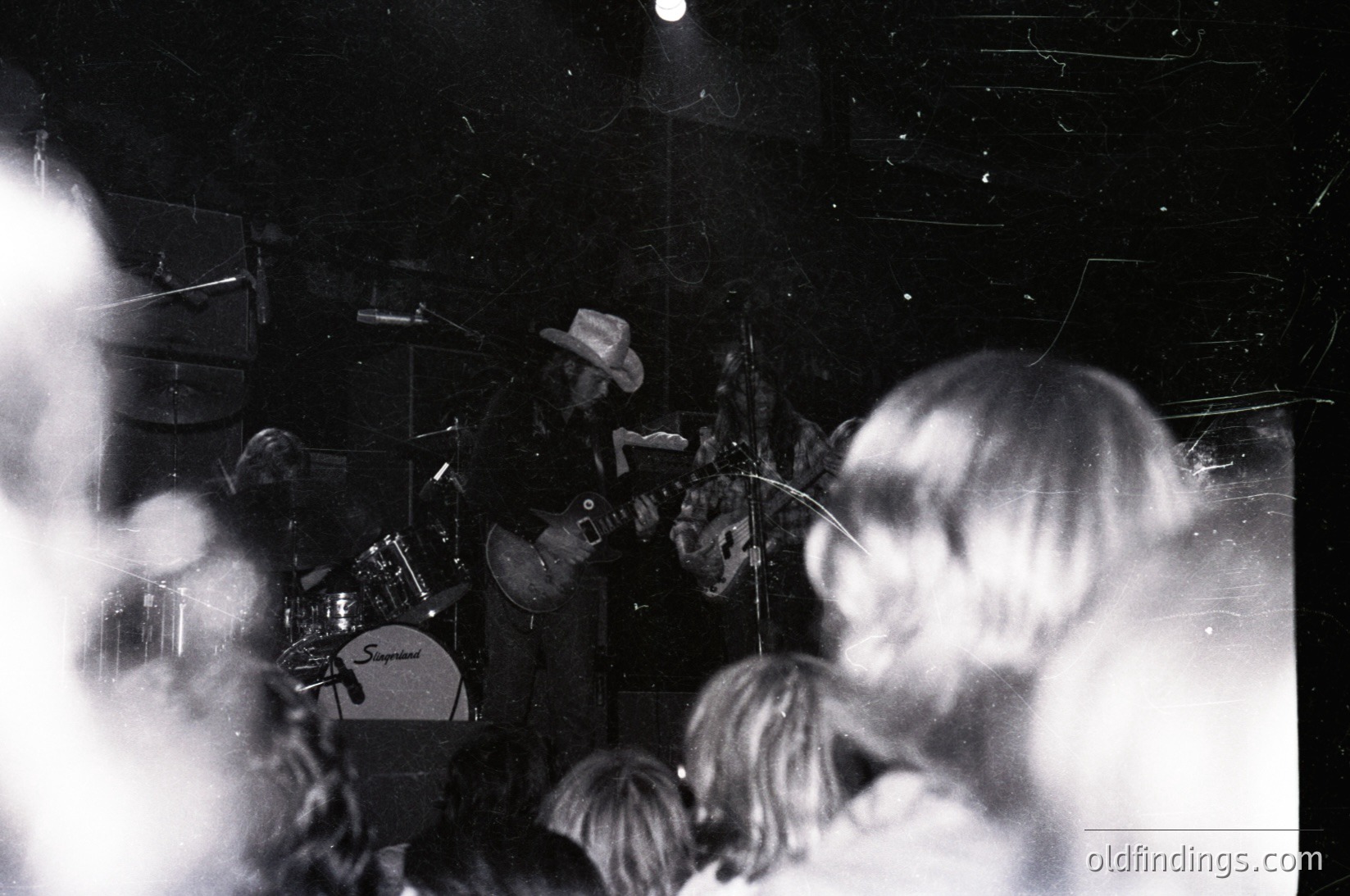 Vintage black-and-white concert shot featuring a guitarist in a cowboy hat playing electric guitar on stage. Crowd silhouetted in foreground, smoke haze obscuring details. Stage setup includes drums and lighting rig. Likely 1960s–1970s live music era.