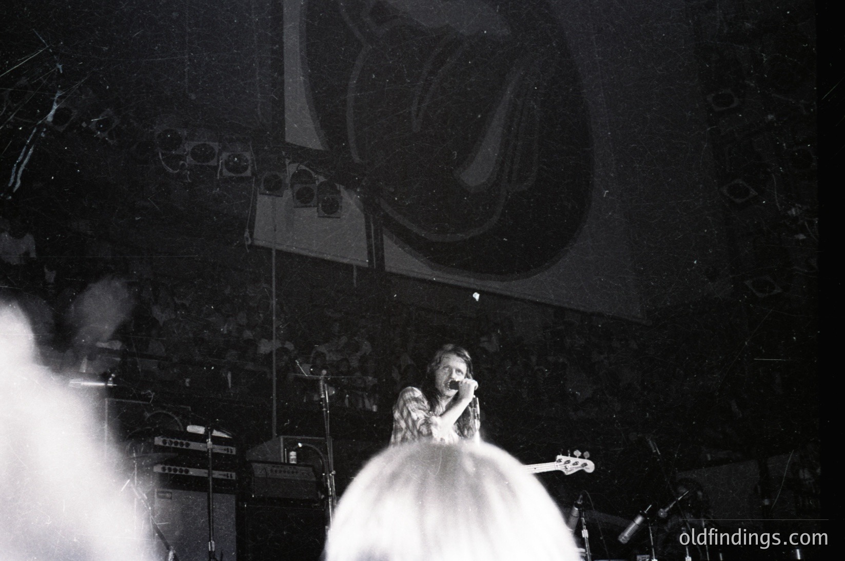 Black-and-white concert shot featuring a vocalist in a flowing white dress, microphone in hand, under dramatic stage lighting. Stage backdrop includes a large, abstract logo and vintage amplifiers. Likely 1970s–1980s rock/psychedelic era.
