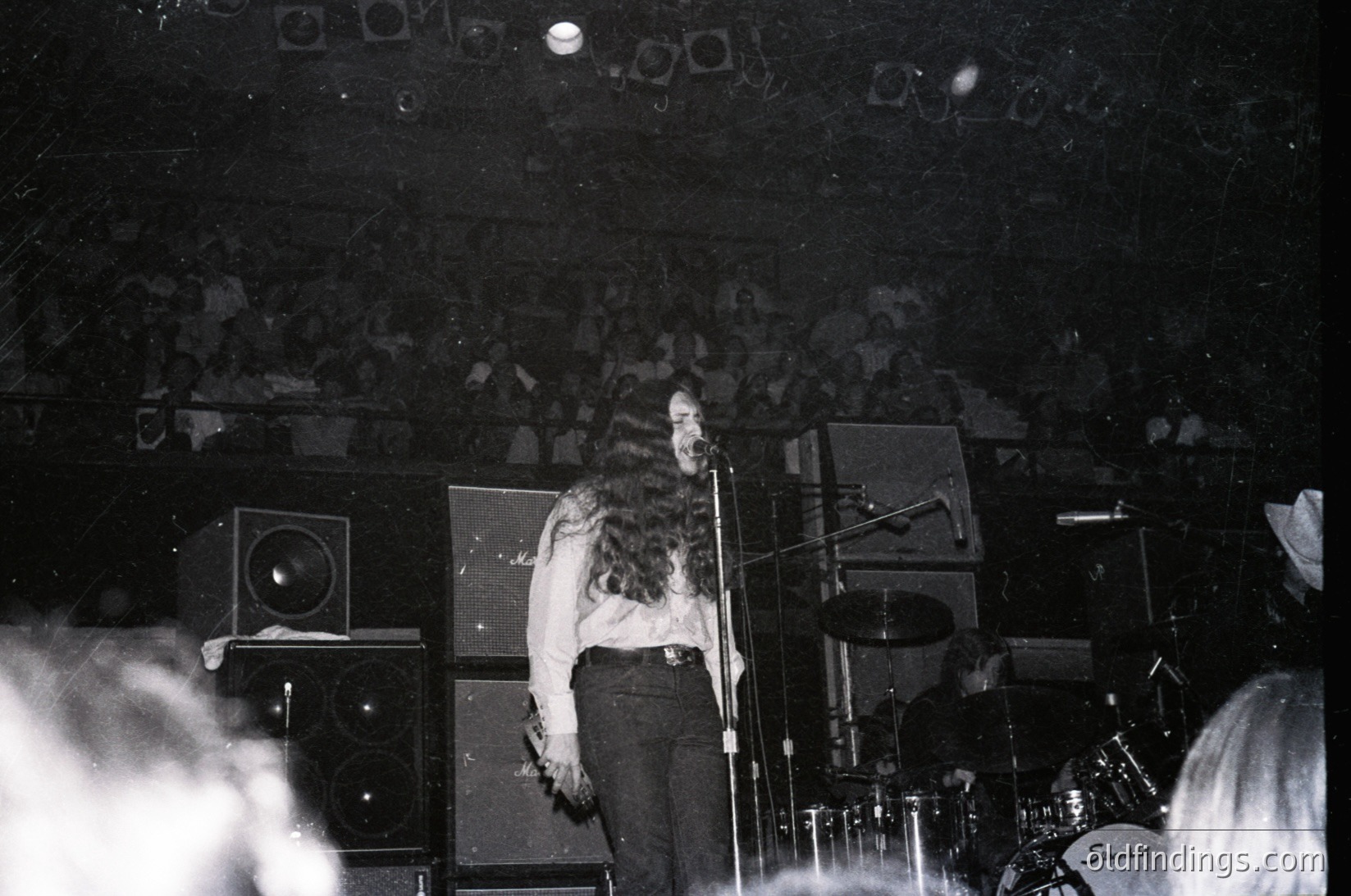 Black-and-white concert shot: musician with curly hair performing on stage, microphone in hand, flanked by vintage amplifiers and a drum kit. Crowd in dimly lit venue, suggesting intimate live music setting. Style evokes 1970s–1980s rock or punk era.