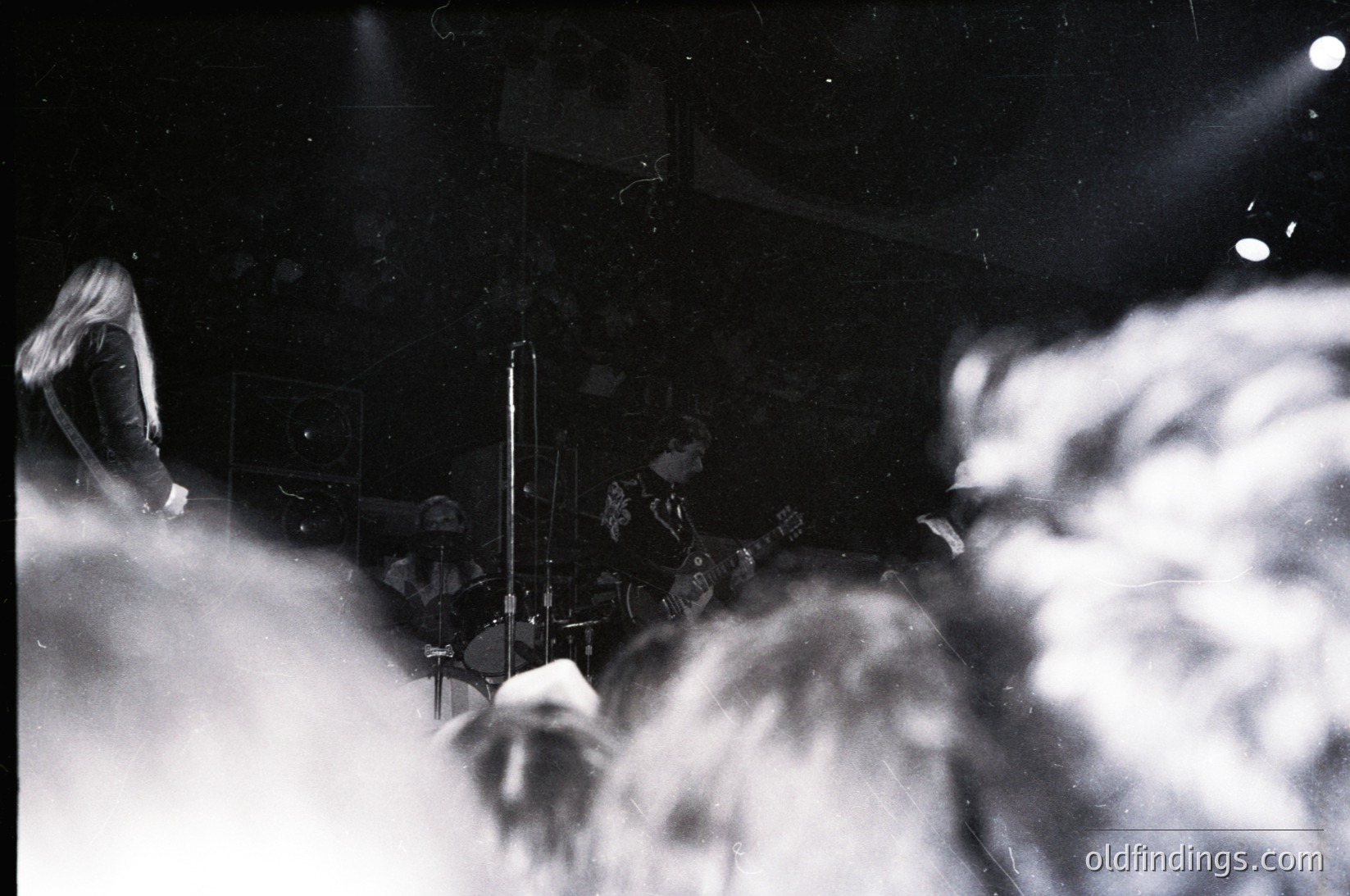 Blurred black-and-white shot of a live jazz band in dimly lit venue, featuring saxophonist and drummer. Crowd silhouetted in foreground, spotlight illuminating performers. Mid-20th century concert atmosphere.