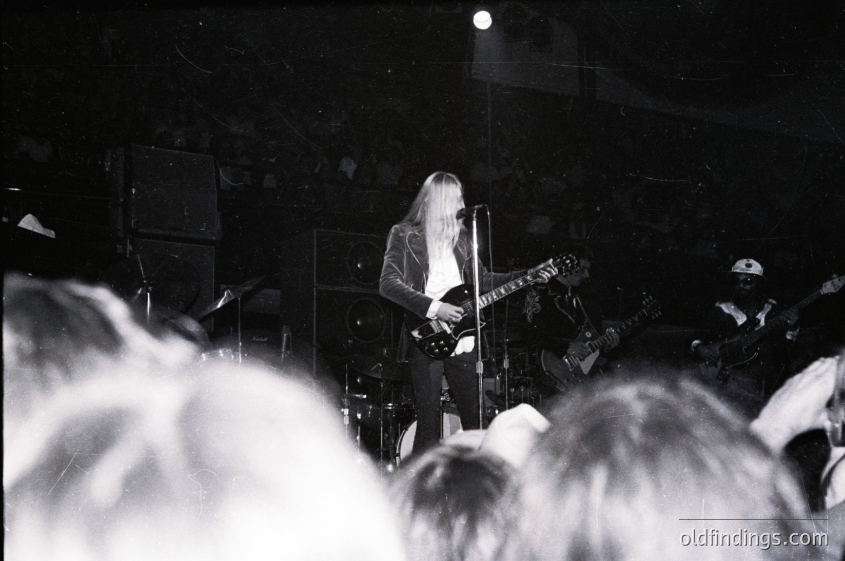 Intimate live performance shot of a guitarist on stage, playing electric guitar with long hair and layered clothing. Crowd silhouetted in foreground, dimly lit venue with visible amplifiers and speakers. Black-and-white, high-contrast style suggests 1970s–1980s rock scene.