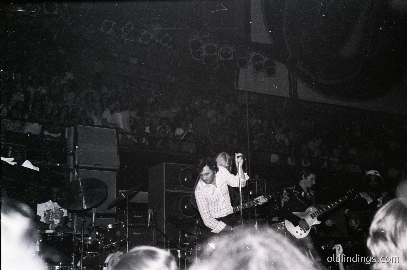Vintage black-and-white concert shot of a guitarist performing onstage, framed by vintage amplifiers and a packed crowd. Stage lighting and grainy film evoke 1960s–1970s live music culture. Crowd silhouettes and blurred motion suggest energetic live performance.