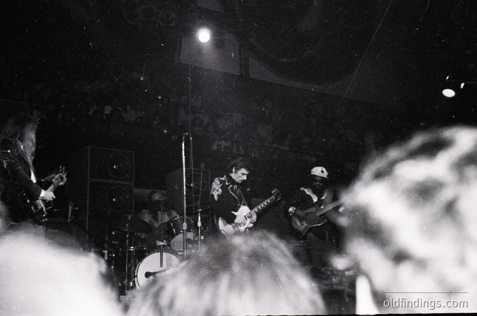 Vintage black-and-white live band performance in dimly lit venue, featuring guitarist, drummer, and vocalist. Stage lighting highlights instruments and audience silhouettes. Mid-20th century concert atmosphere with analog audio equipment visible. Ideal for music history, vintage photography, and live event archives.