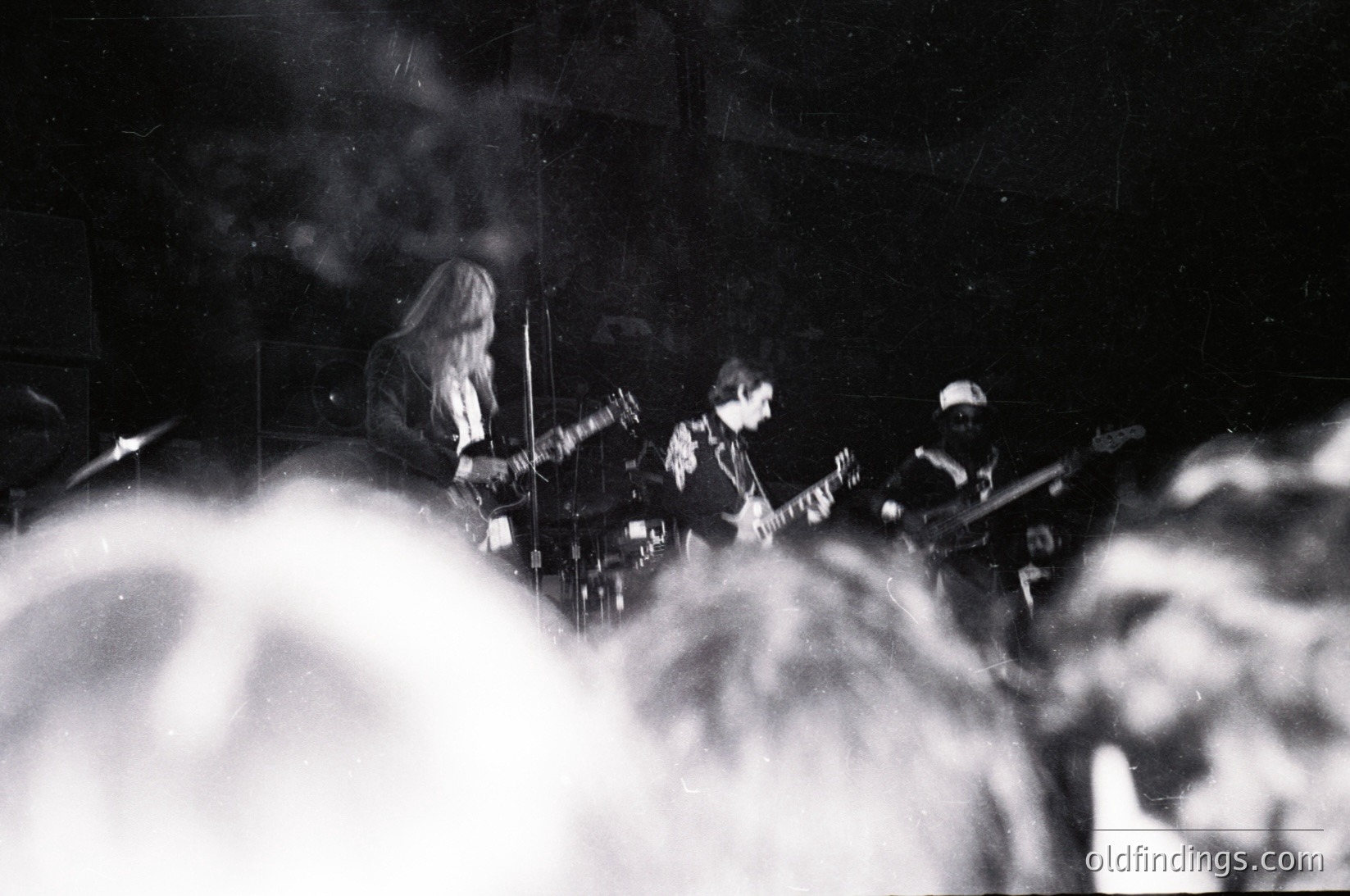 Black-and-white live band performance with dense stage smoke. Three musicians—guitarist (center), bassist (left), and drummer (right)—immersed in performance. Long hair and vintage attire suggest 1960s–1970s rock era. Intimate, atmospheric stage lighting enhances dramatic effect.