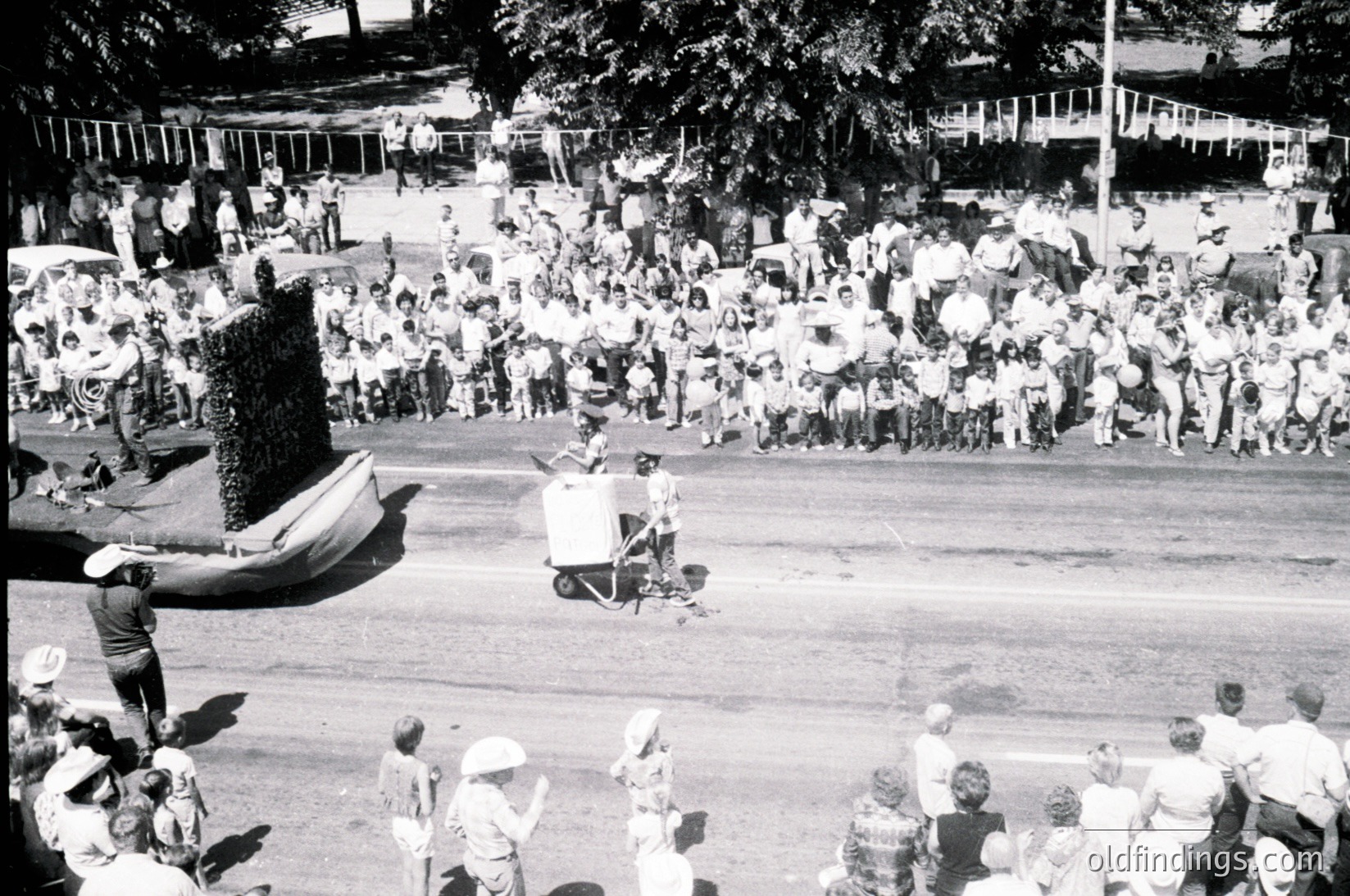 Aerial shot of a mid-20th-century outdoor event featuring a large crowd in casual summer attire. Central figure in a white suit and hat performs on a small stage with a microphone, surrounded by a decorated structure resembling a giant cake or sculpture. Trees and parked cars suggest a public park or plaza setting.