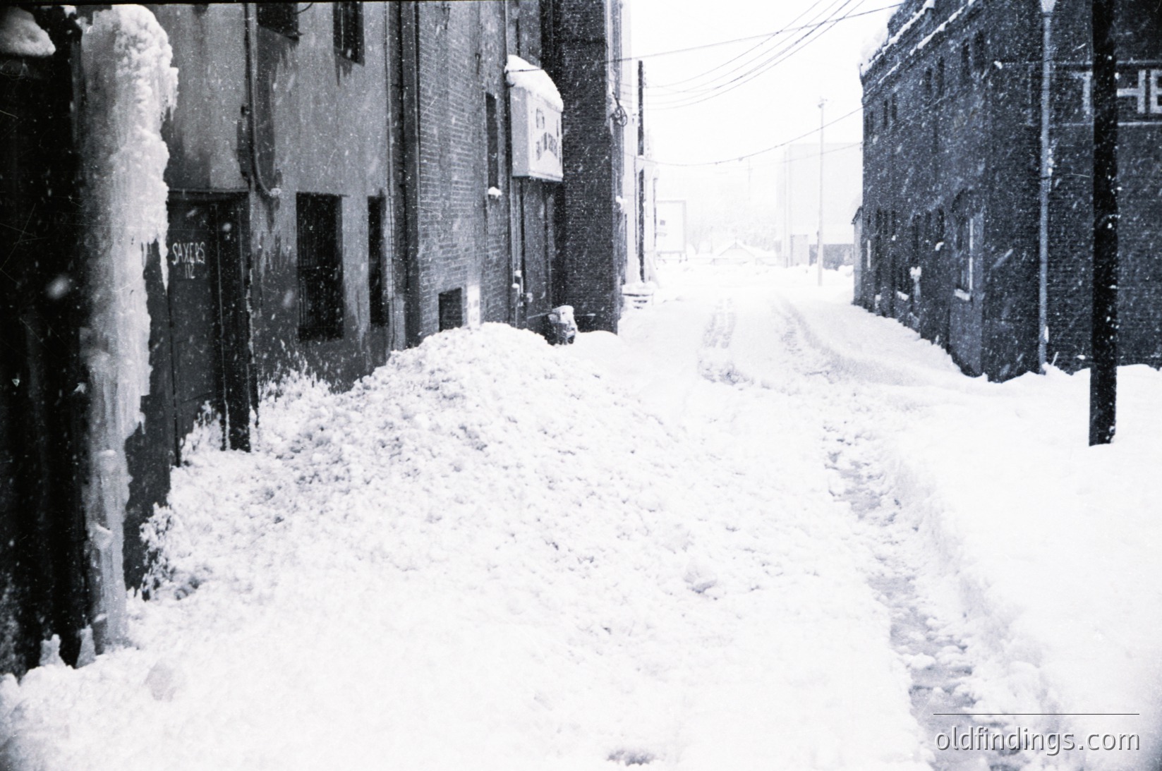 Blizzard-covered urban alleyway with heavy snow accumulation on buildings and sidewalks, likely mid-20th century. Snowdrifts obscure street signs and pedestrian paths, emphasizing isolation. Architectural details include brick facades and a visible "Savings" sign.
