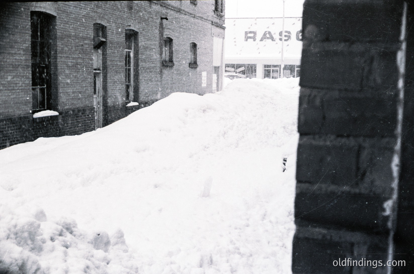 Mid-20th century urban winter scene: snow-covered brick buildings with boarded windows, likely post-WWII reconstruction. Prominent signage "RAS" suggests industrial or commercial use. Graffiti-like "2" on snow hints at playful or temporary marking. Black-and-white grain adds vintage authenticity.