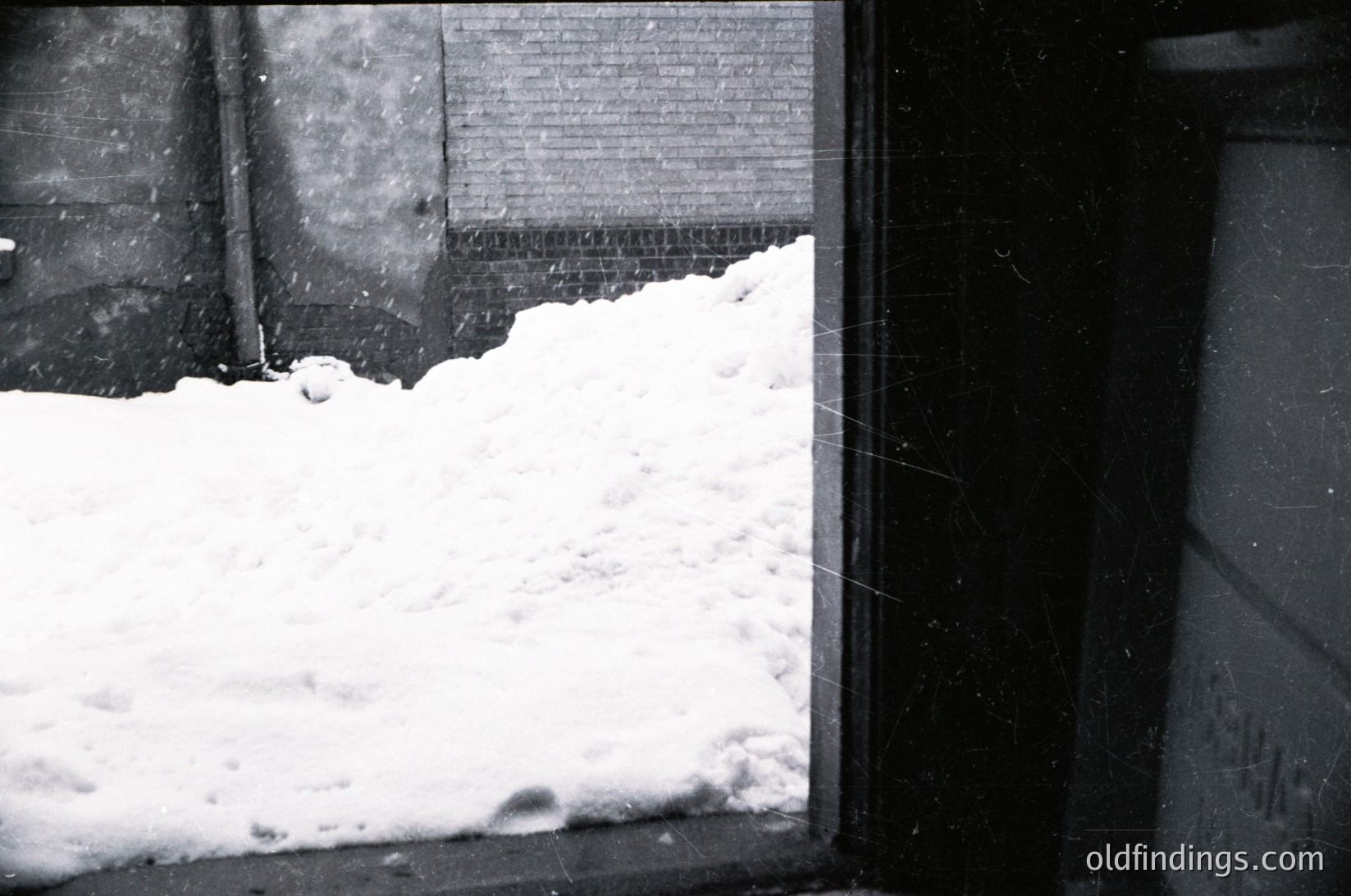 Black-and-white urban winter scene: snow piled against a brick wall and window frame, suggesting heavy snowfall. Industrial pipe and metal door frame visible. Likely mid-20th century due to architectural style and monochrome.