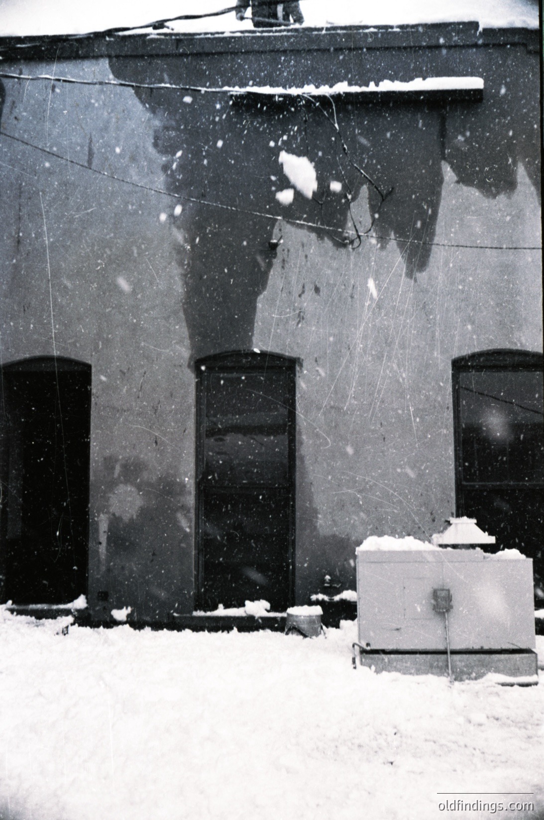 Snow-covered urban facade with three tall, narrow windows framed by worn concrete. Overhead wires and a utility box with snow accumulation. Mid-century Soviet-era architecture, likely Eastern Bloc.