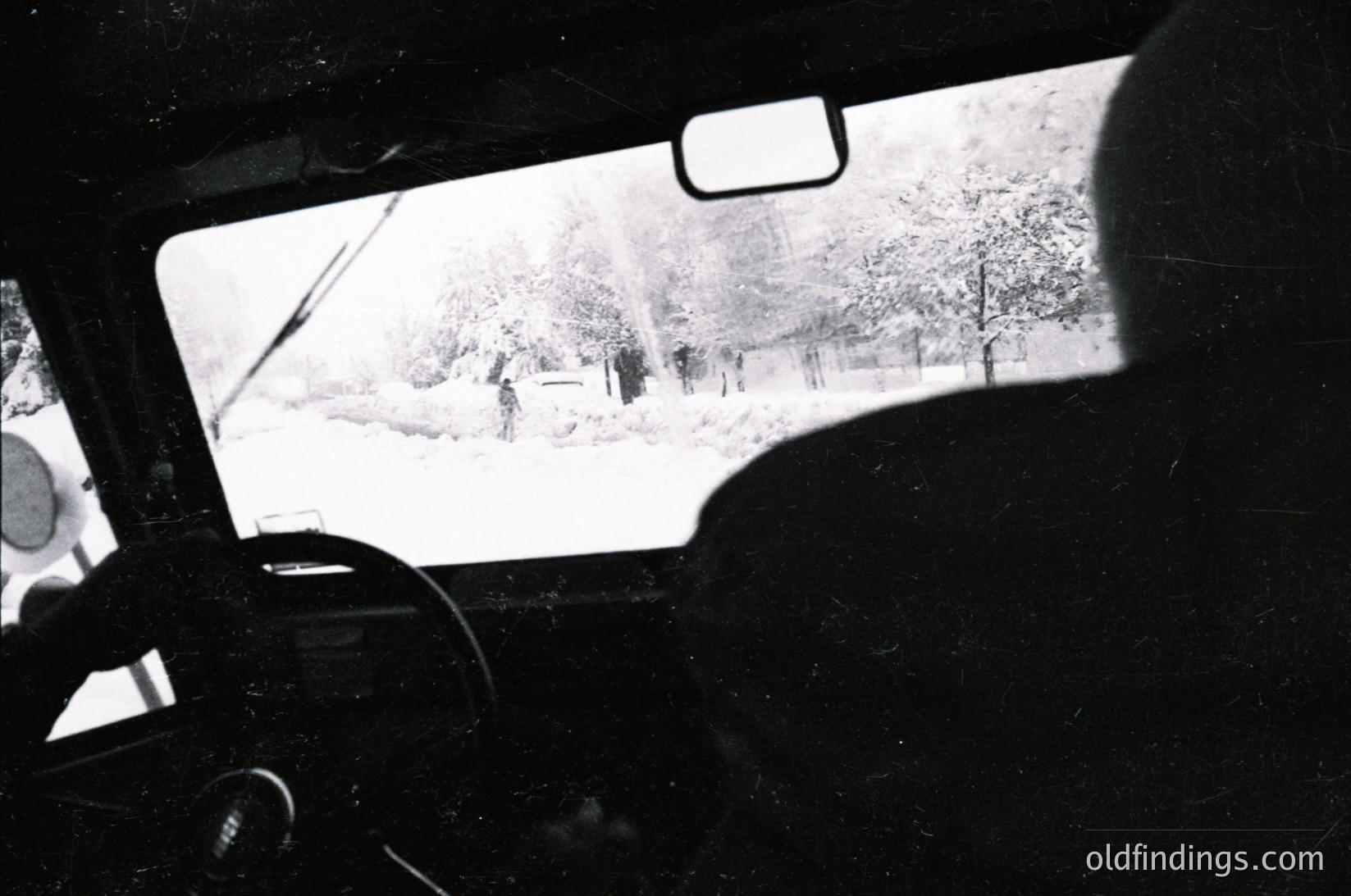 Black-and-white shot from inside a vintage car, snow-covered landscape visible through windshield. Distant figures in winter attire walking along a snow-lined path. Classic winter scene evokes nostalgia.