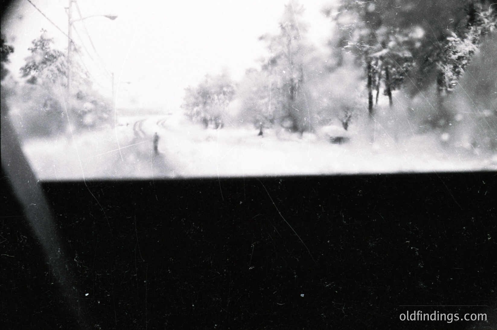 Blurred monochrome shot of a snowy winter scene viewed through a windowpane, showing indistinct figures walking on a snow-covered path flanked by leafless trees. Vintage grain and frost patterns on glass add texture. Likely mid-20th century European countryside.
