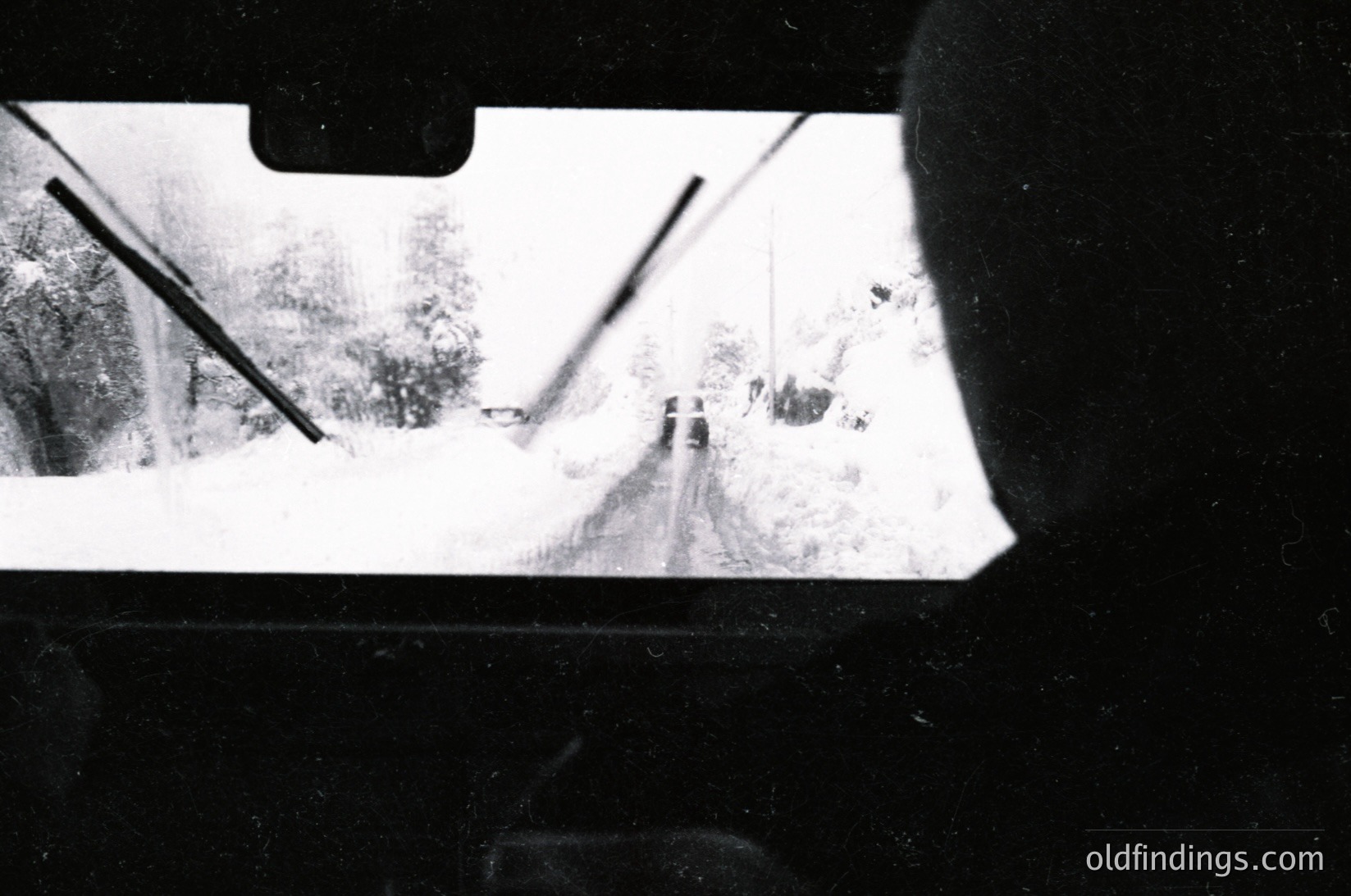 Blurred rearview shot of a snow-covered road with minimalist composition—bare trees, faint tire tracks, and a lone figure in distance. Black-and-white, high-contrast aesthetic evokes mid-20th century travel or documentary photography.