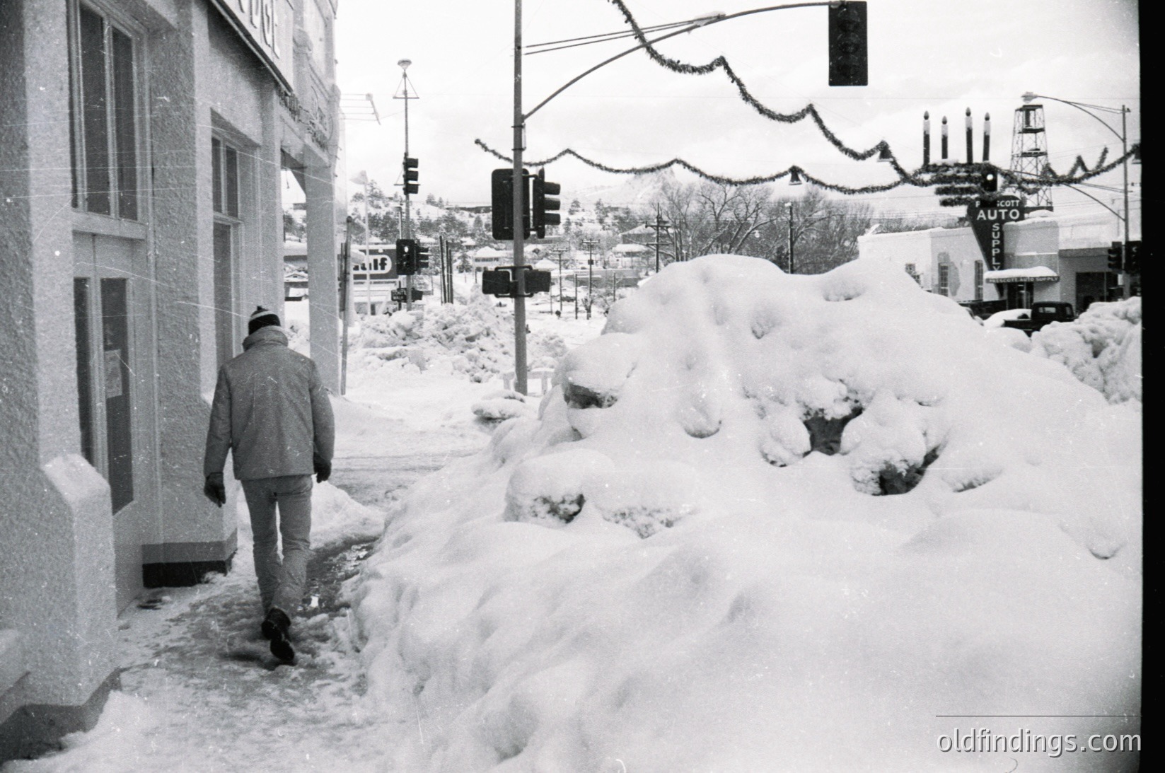 Black-and-white street scene featuring a lone man in winter attire navigating a snow-covered sidewalk. Snowbanks line the road, obscuring traffic signals and streetlights. Decorative holiday garlands stretch overhead. Mid-century urban architecture with brick buildings and industrial elements in background. Likely or North American city.