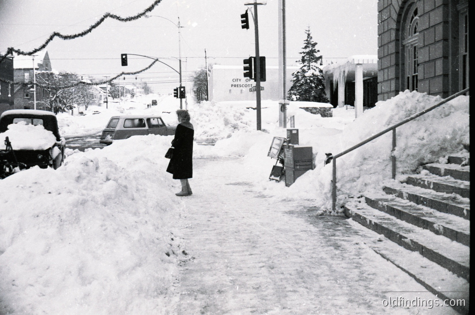 Mid-20th century winter street scene in Prescott, AZ. Woman in winter coat navigates snow-covered sidewalk beside vintage cars and a snowbanked building. Historic downtown architecture with "City of Prescott" sign visible.