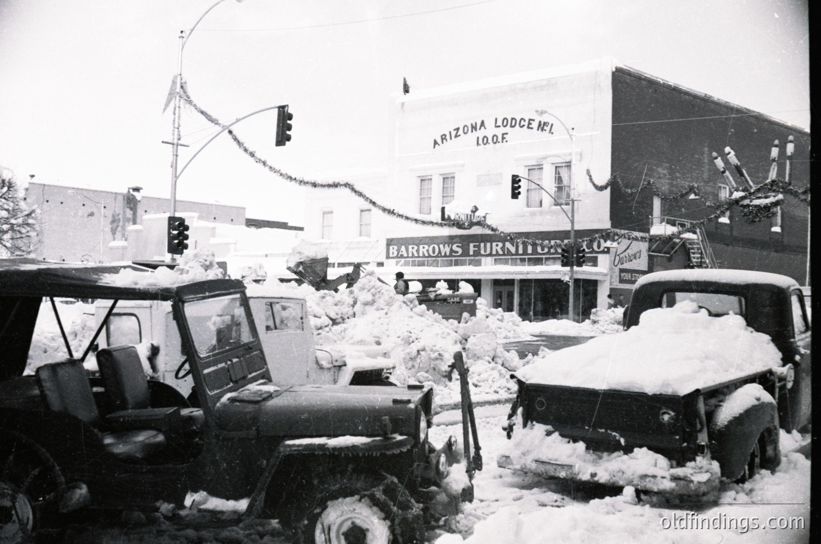 Mid-20th century snowstorm disrupts downtown streets, with deep drifts covering vehicles and sidewalks. Snow-laden traffic lights and a partially buried jeep dominate the foreground, while businesses like "Arizona Lodge" and "Barrows Furniture" signage remain visible. Snow-covered signage and holiday lights hint at winter holiday season. Classic 1950s-60s urban landscape.