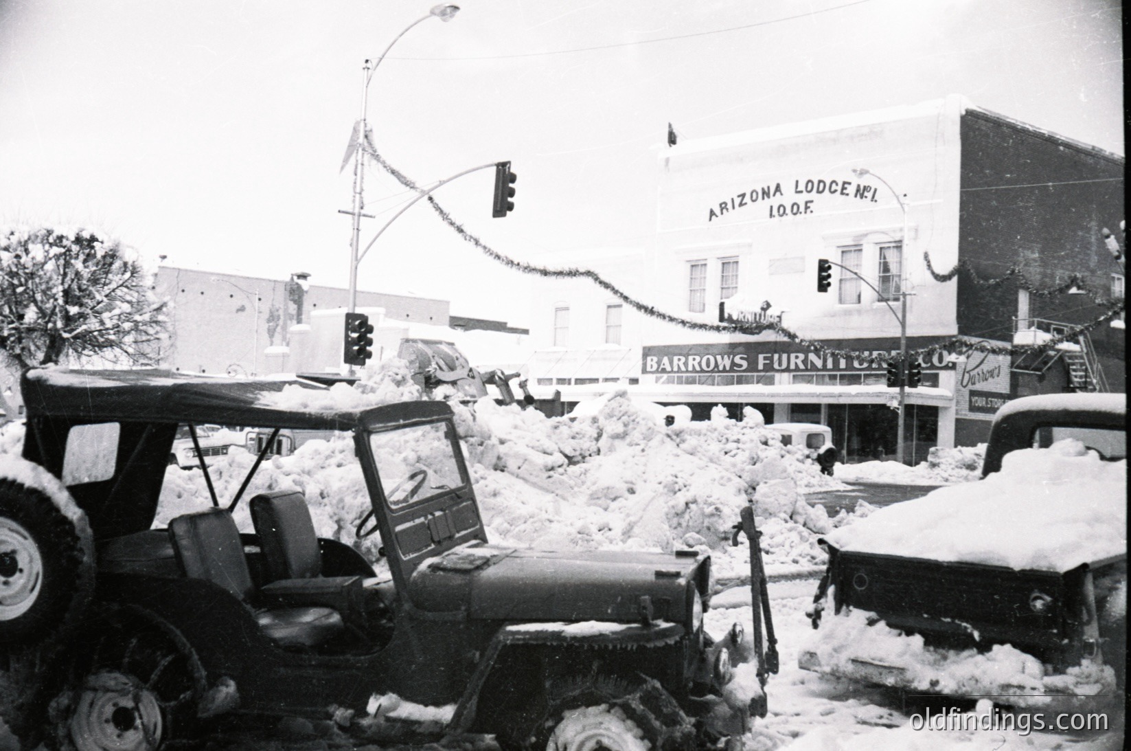 Mid-20th century street scene after heavy snowfall, featuring a tractor clearing snow in downtown. Visible: "Arizona Lodging" sign (10th floor) and "Barrows Furniture" storefront. Snow-covered vehicles and traffic lights indicate urban infrastructure. Likely --- *Note: Time period inferred from architectural style, vehicle design, and signage.*