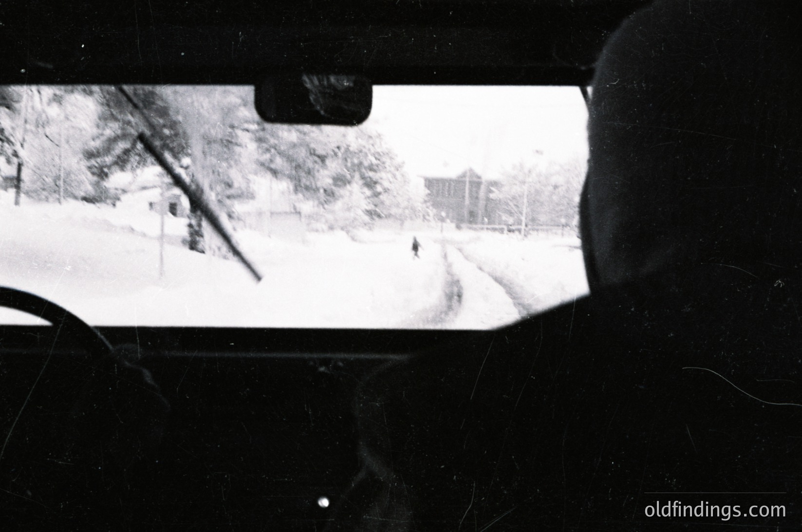 Black-and-white shot from inside a vehicle, framed by windshield and rearview mirror. Snow blankets a residential street with leafless trees and distant buildings, suggesting winter conditions. Composition captures motion blur from driving, evoking a sense of travel.
