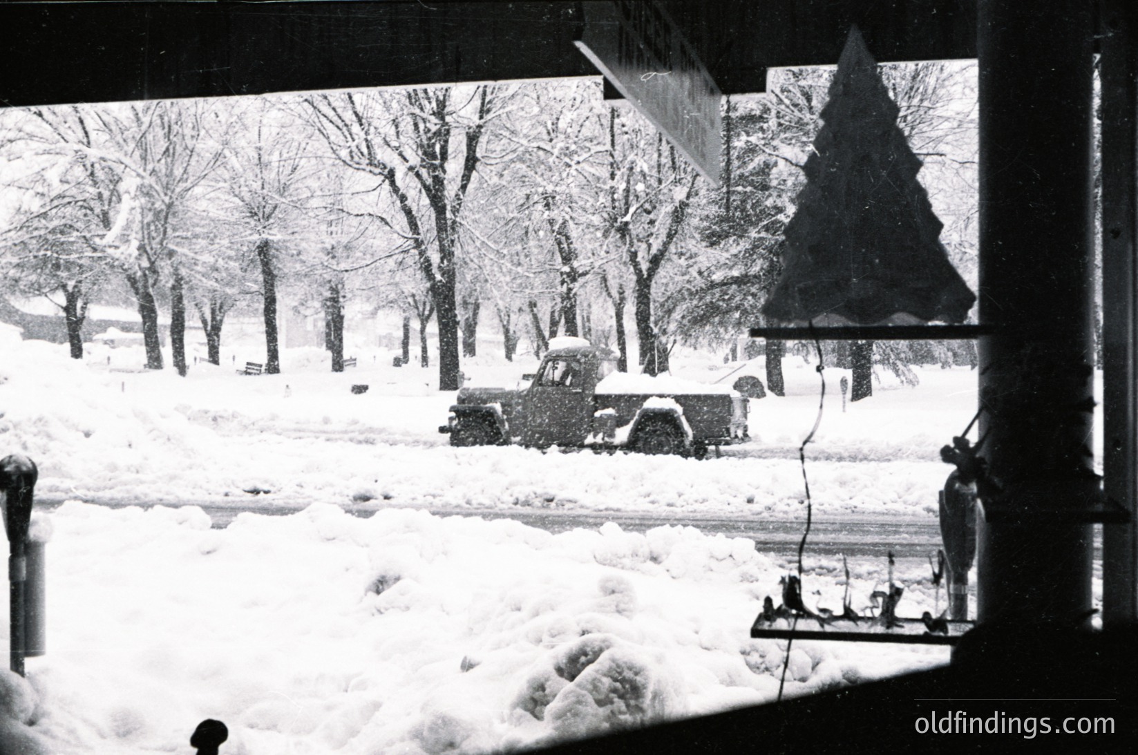 Vintage black-and-white winter scene showing a snow-covered urban park. Snow-laden vintage truck parked near leafless trees, with a triangular Christmas tree decoration visible. Snowdrifts obscure pathways, and a person in winter attire shovels snow near a fence. Indoor window frame frames the view, suggesting a residential interior. Likely mid-20th century () in a temperate climate city.