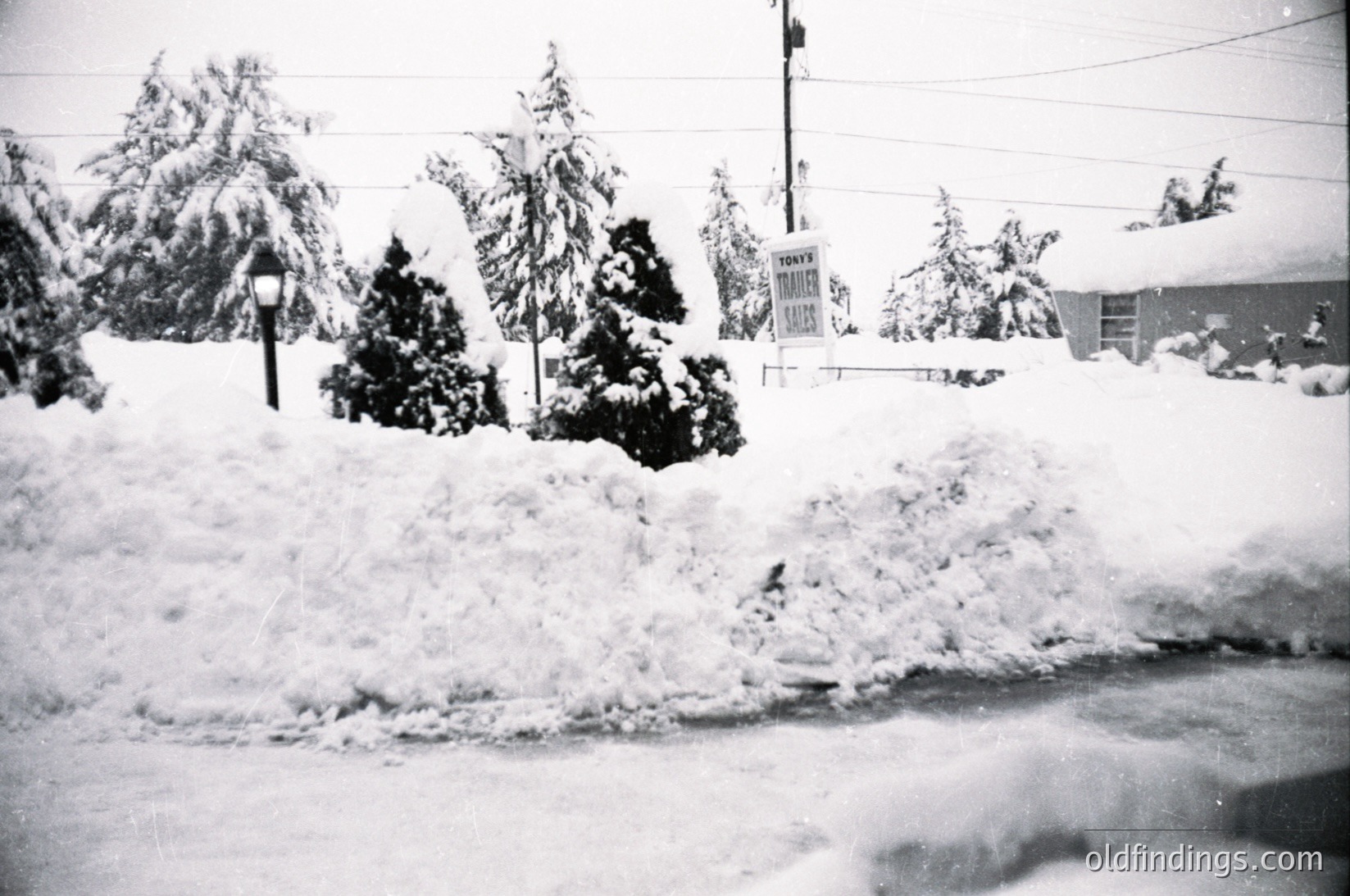 Black-and-white street scene blanketed in heavy snow, likely mid-20th century. Snow-covered evergreens, utility poles, and a partially visible "TOWNY" sign suggest a suburban or small-town setting. Snowbanks line the road, obscuring sidewalks.