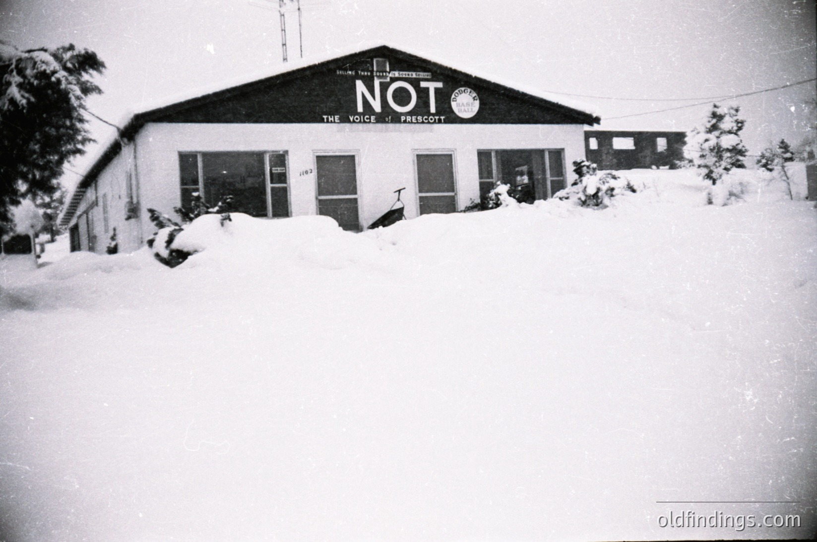 Black-and-white photo of a snow-covered building labeled **"NOT: The Voice of Prescott"**—likely a 1960s–1970s radio station in Prescott, AZ. Snow blankets the ground, windows, and roof, with sparse evergreen trees framing the scene. Simple, utilitarian architecture with large windows and a signboard.