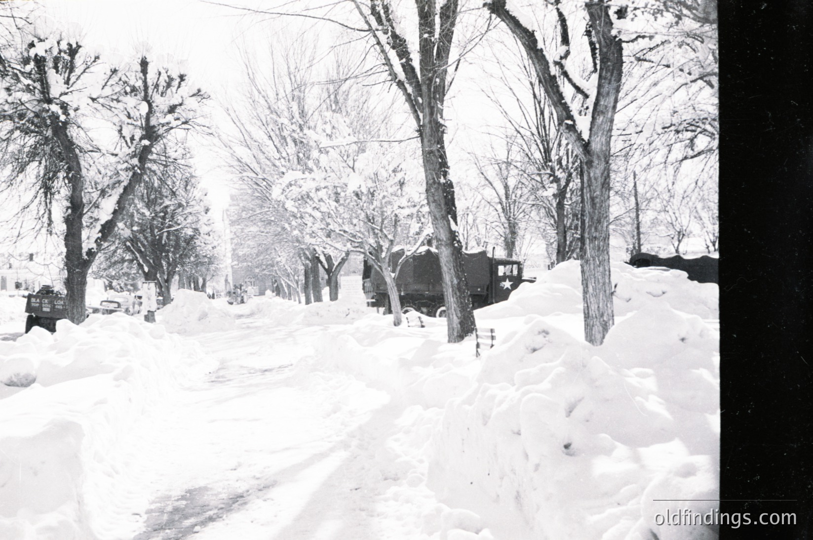 Mid-20th century winter street scene with deep snow covering a residential road. Bare trees line both sides, and a vintage car is parked near a house with a dark, sloped roof. Snowbanks and tire tracks mark the path. Likely urban/suburban setting, --- *Note: Time period inferred from car design and photographic style.*