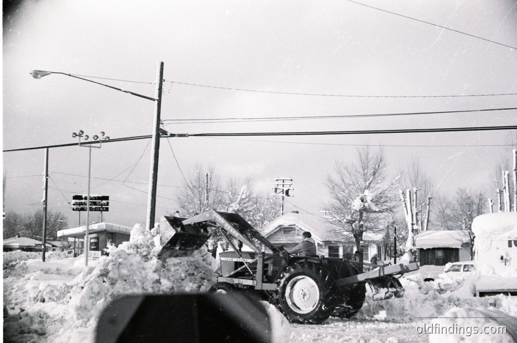 Mid-20th century winter street scene with heavy snow accumulation. Snow-covered utility truck and parked cars on a residential road, flanked by leafless trees and utility poles. Overhead wires and street signs suggest urban/suburban setting.