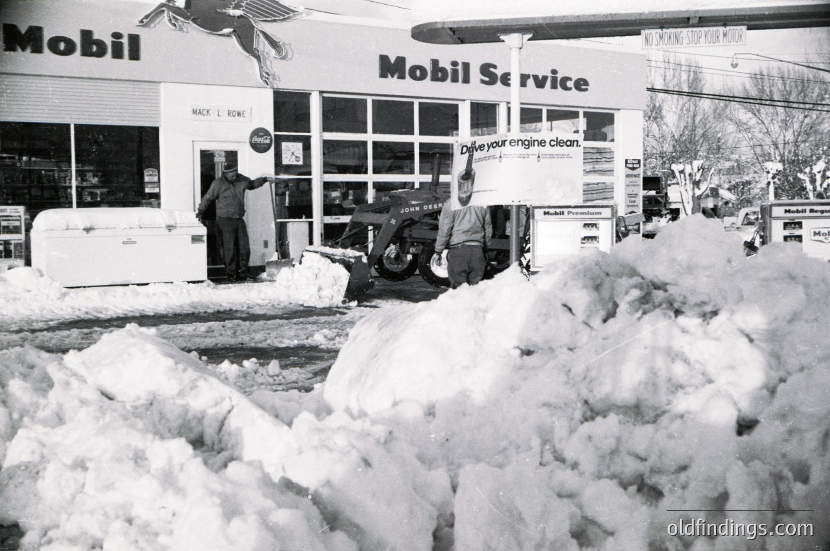Mid-20th century Mobil Service station buried under heavy snowfall. Two workers clear snow with a snowblower and shovel near entrance. Signage includes "Drive your engine clean" and "Mack L. Rowe" auto parts. Snowdrifts block visibility of surrounding area, emphasizing winter conditions.