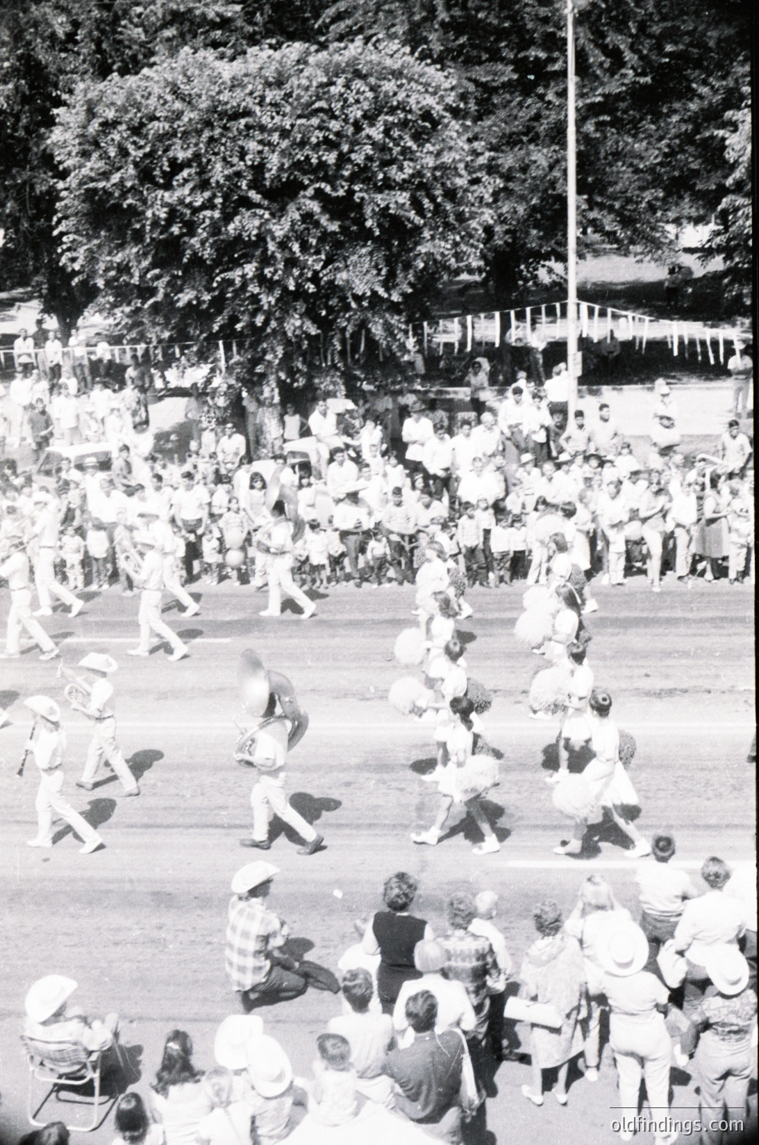Aerial shot of a lively outdoor festival or parade, likely from the **1960s–1970s**. Crowds gather along a tree-lined street, watching participants in coordinated costumes (skirted outfits, hats) perform a choreographed routine. Decorative banners and a flagpole suggest a formal or celebratory event. Urban setting with visible architecture and spectators seated/standing.