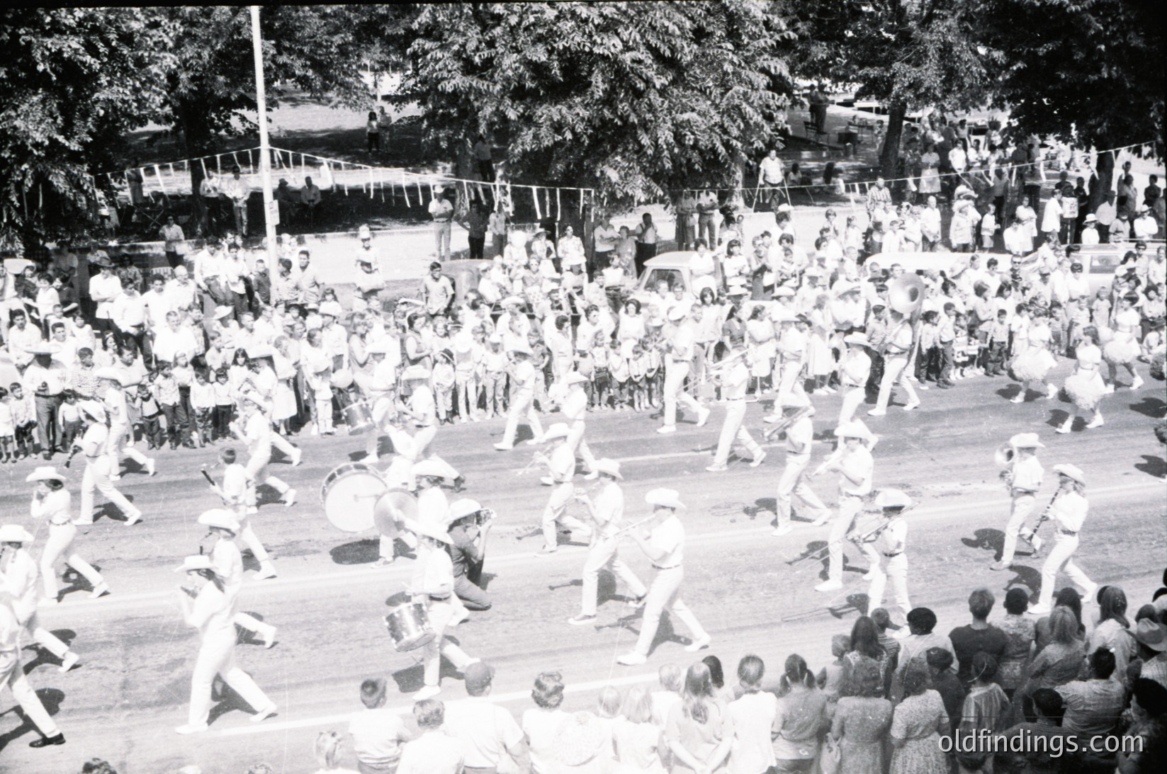 Crowded outdoor dance event featuring energetic group dancing in a circular formation, likely a folk or traditional celebration. Mid-20th century attire (1950s–1960s) with men in suits and women in dresses. Spectators line the perimeter, some seated on grass. Trees and parked vehicles suggest a park or public square setting.