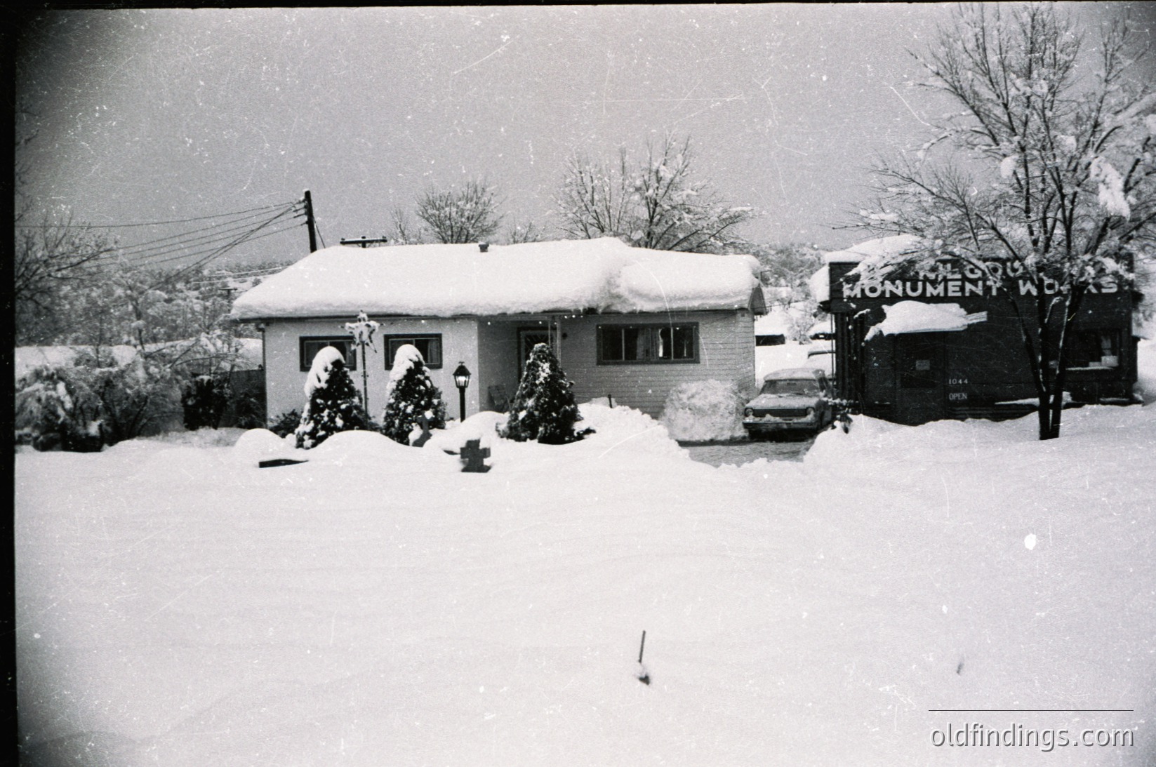 Mid-century suburban home blanketed in snow, featuring a single-story ranch-style design with arched windows. Decorated with two Christmas trees in the front yard. "Monument Works" sign visible in background, suggesting local business presence. Likely 1950s–1960s USA.