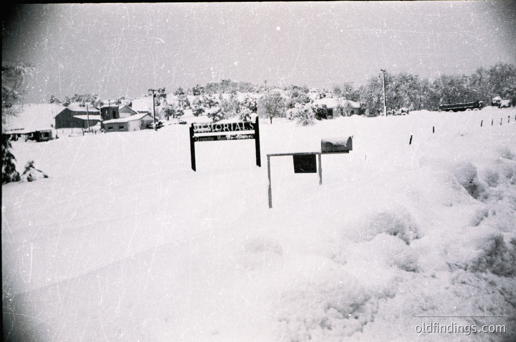Mid-century rural landscape buried under heavy snowfall, likely 1950s–1960s. Snow-covered farmhouses, utility poles, and a wooden mailbox dominate the scene. Dense forest in background suggests a colder climate region.