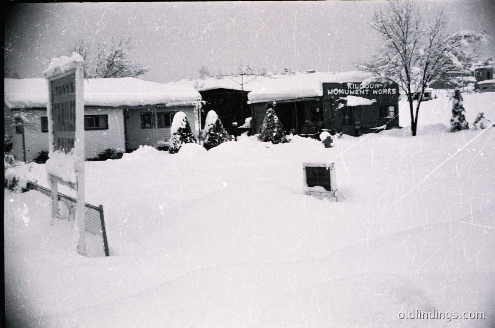 Mid-20th century snowy street scene with light industrial/commercial buildings. Snow-covered signage reads "Toyes" and "Monument Works." Bare trees and parked vehicles suggest winter conditions. Likely U.S. small-town setting, 1950s–1960s.