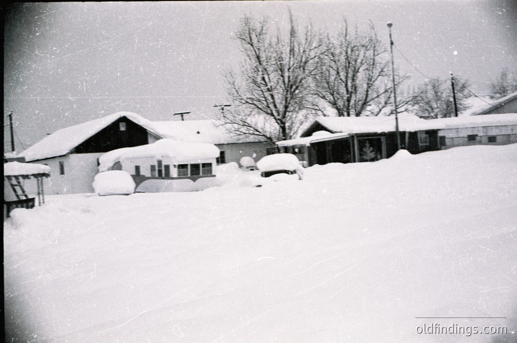 Mid-century suburban street blanketed in heavy snow, likely 1950s–1960s. Single-story homes with gabled roofs, narrow driveways, and parked cars partially buried. Bare trees and utility poles frame the scene. Classic American postwar residential architecture.