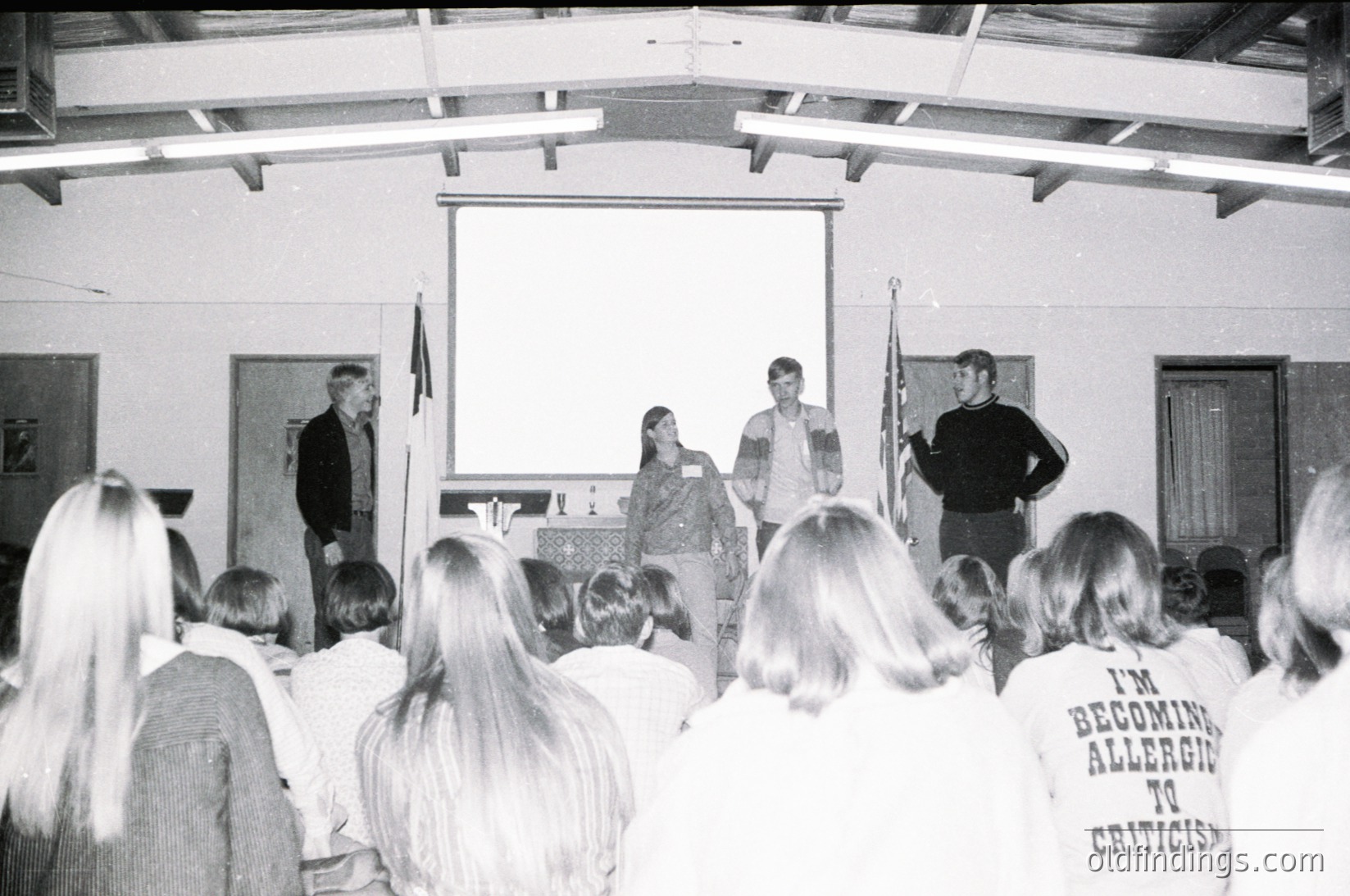 Black-and-white indoor lecture hall scene with four speakers at a podium, facing an attentive audience. Visible text on a T-shirt reads *"I'm becoming allergic to criticism."* Architectural details include a curved ceiling and a flag on the right. Likely 1970s–1980s based on fashion and photography style.
