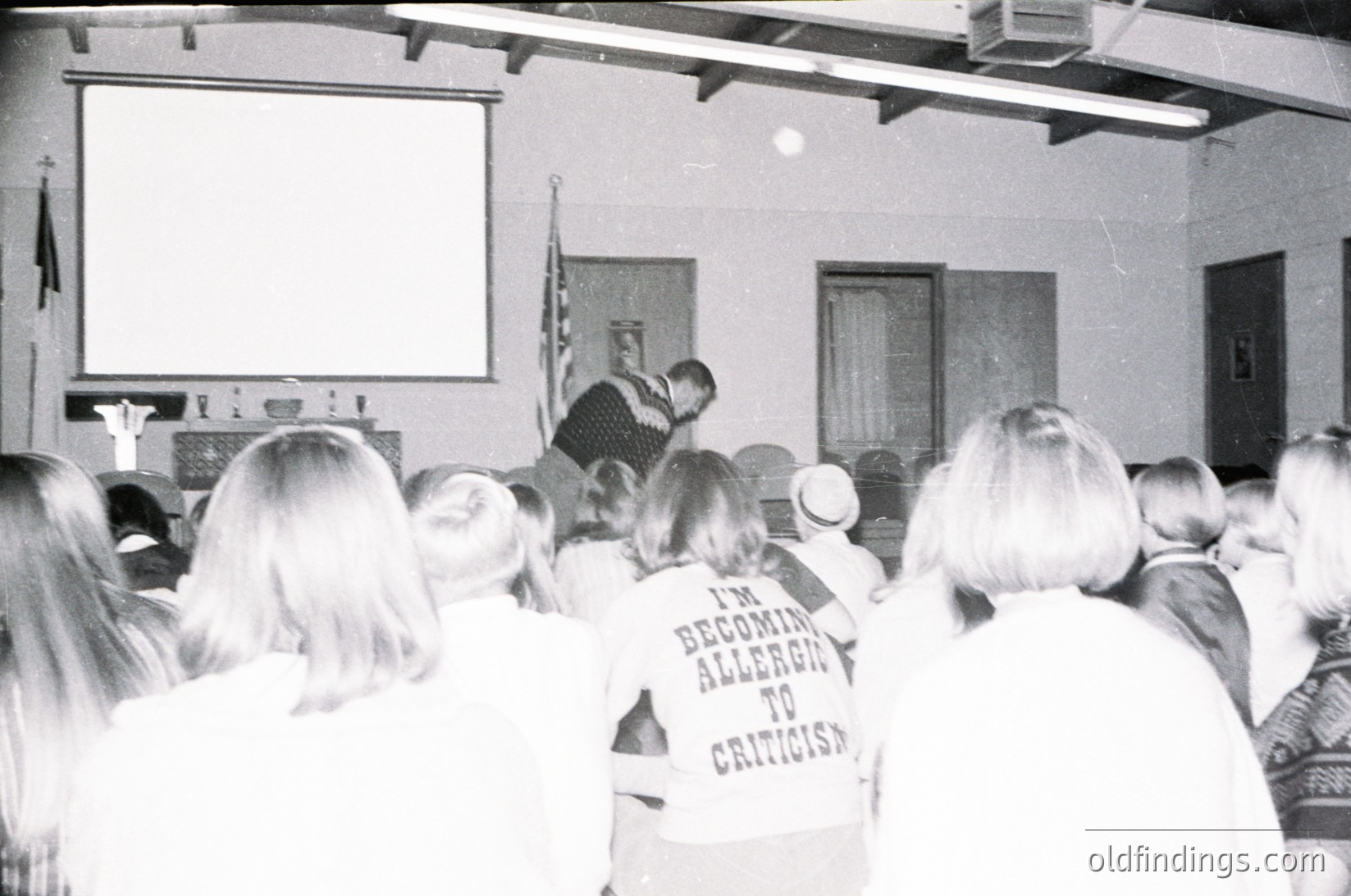 1970s indoor lecture hall with projector screen. Speaker addresses audience; attendees wear humorous "I'm Becoming Allergic to Criticism" shirts. Simple wooden pews and American flag visible. Likely educational or corporate event.