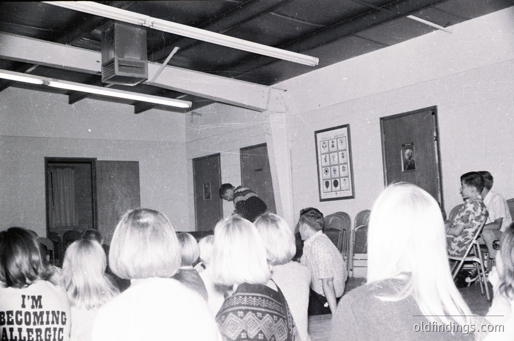 A black-and-white classroom scene from the 1970s–1980s, featuring a group of seated students facing a teacher at a front lectern. The room includes a projector mounted on the ceiling, a bulletin board with geometric patterns, and a door with a framed portrait. Casual, patterned clothing and hairstyles reflect mid-century educational aesthetics.