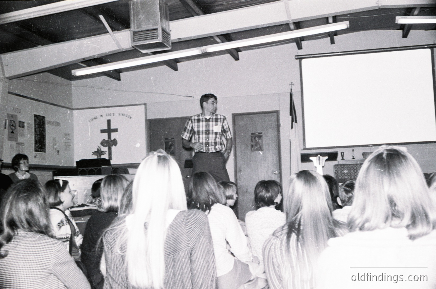 A 1970s classroom scene: teacher in plaid shirt addresses seated students. Cross and "Love and God" banner on wall. Industrial ceiling vents and projector screen visible.