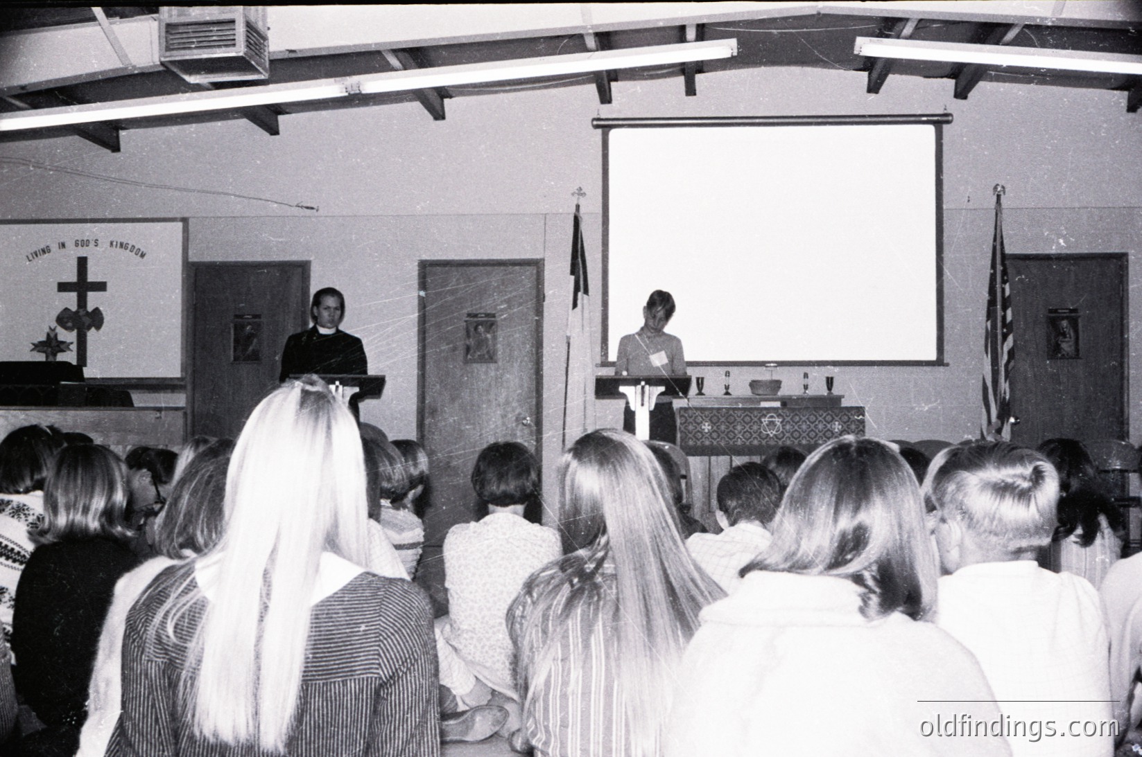 A black-and-white wedding ceremony in a church hall, likely 1970s–1980s. Bride in veil stands at altar with groom beside her; officiant delivers vows at podium. Wooden pews filled with attendees, religious symbols (cross, altar cloth) visible. Formal attire and indoor setting highlight mid-century wedding traditions.