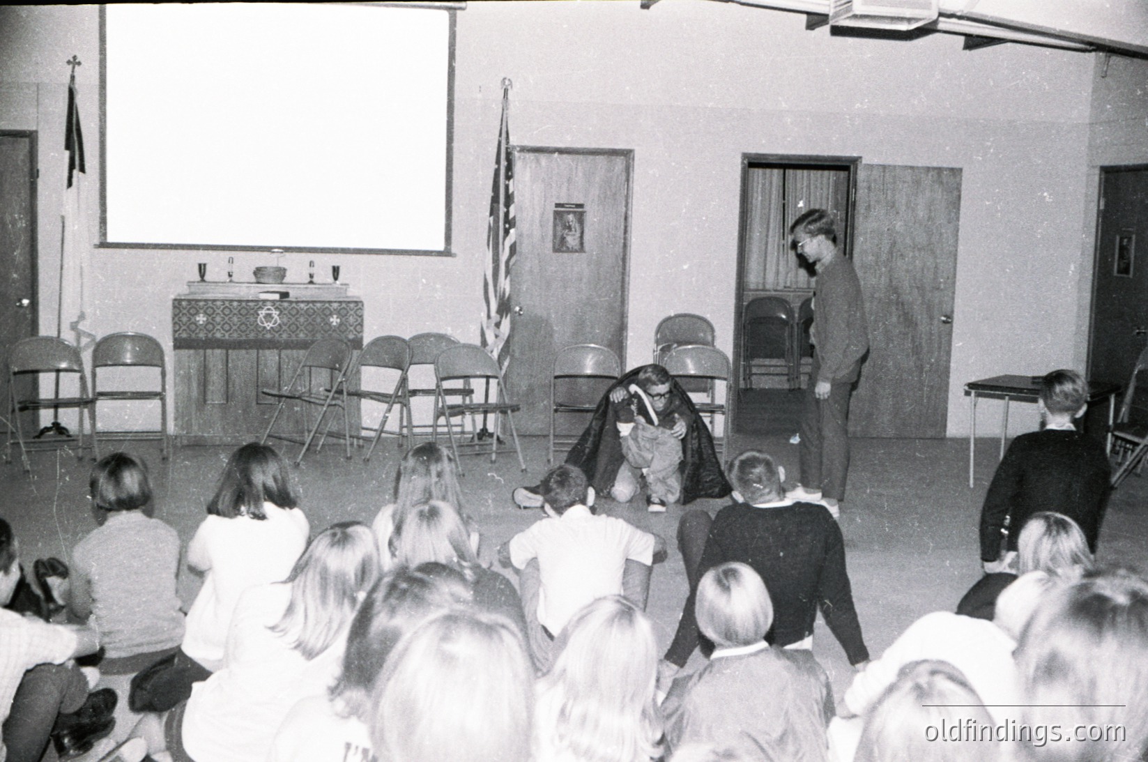 A 1970s-era classroom or assembly hall featuring a puppet show. A performer in costume interacts with a puppet on stage while a crowd of children and adults observe. Stage setup includes a projector screen, wooden chairs, and a lectern with a flag. Formal attire suggests a school or community event.