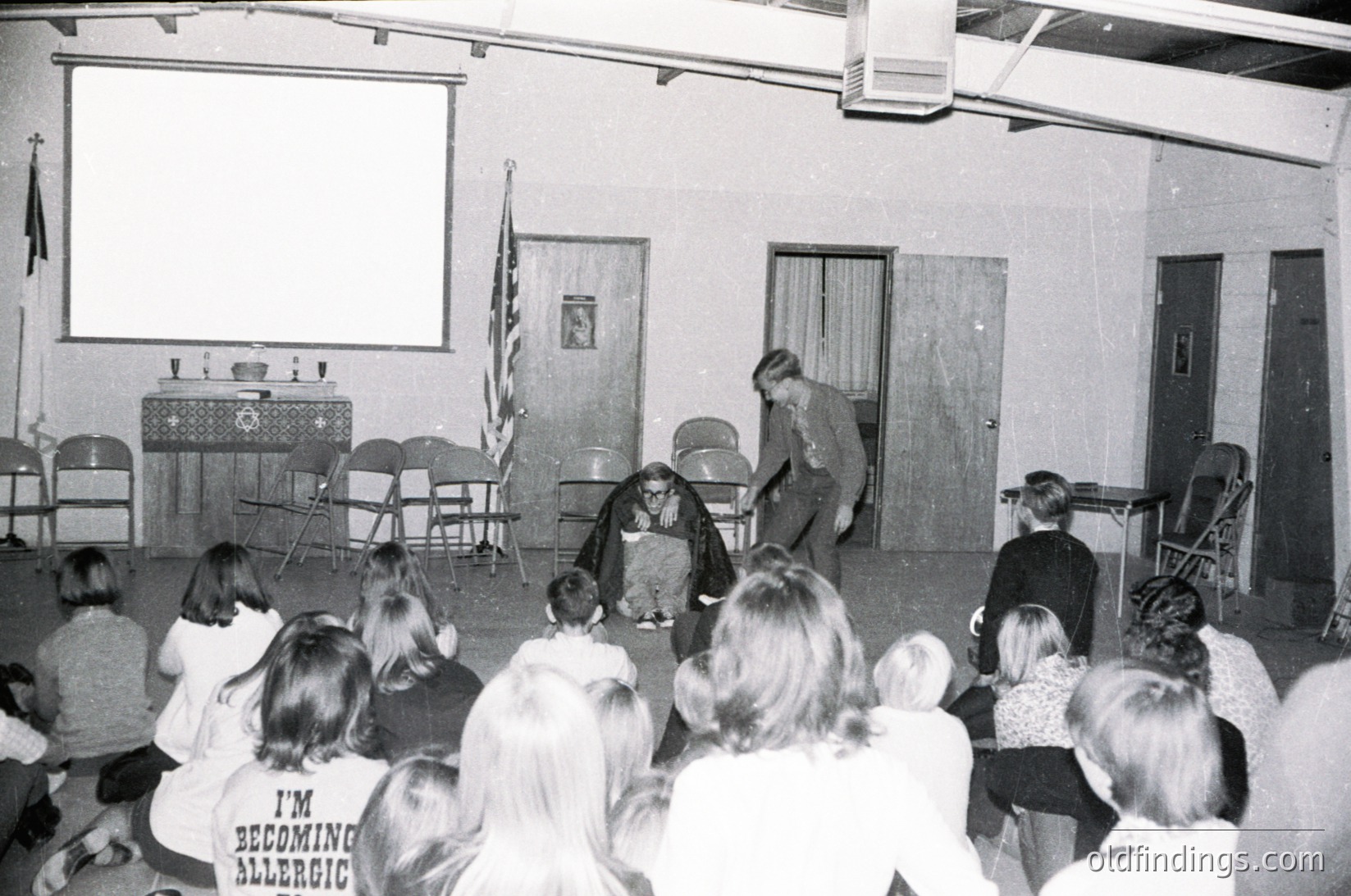 Black-and-white classroom scene featuring a teacher demonstrating science experiment with liquid nitrogen to a seated audience of students. Stage setup includes a projector screen, American flag, and vintage wooden chairs. Mid-20th century educational environment, likely 1960s–1970s.